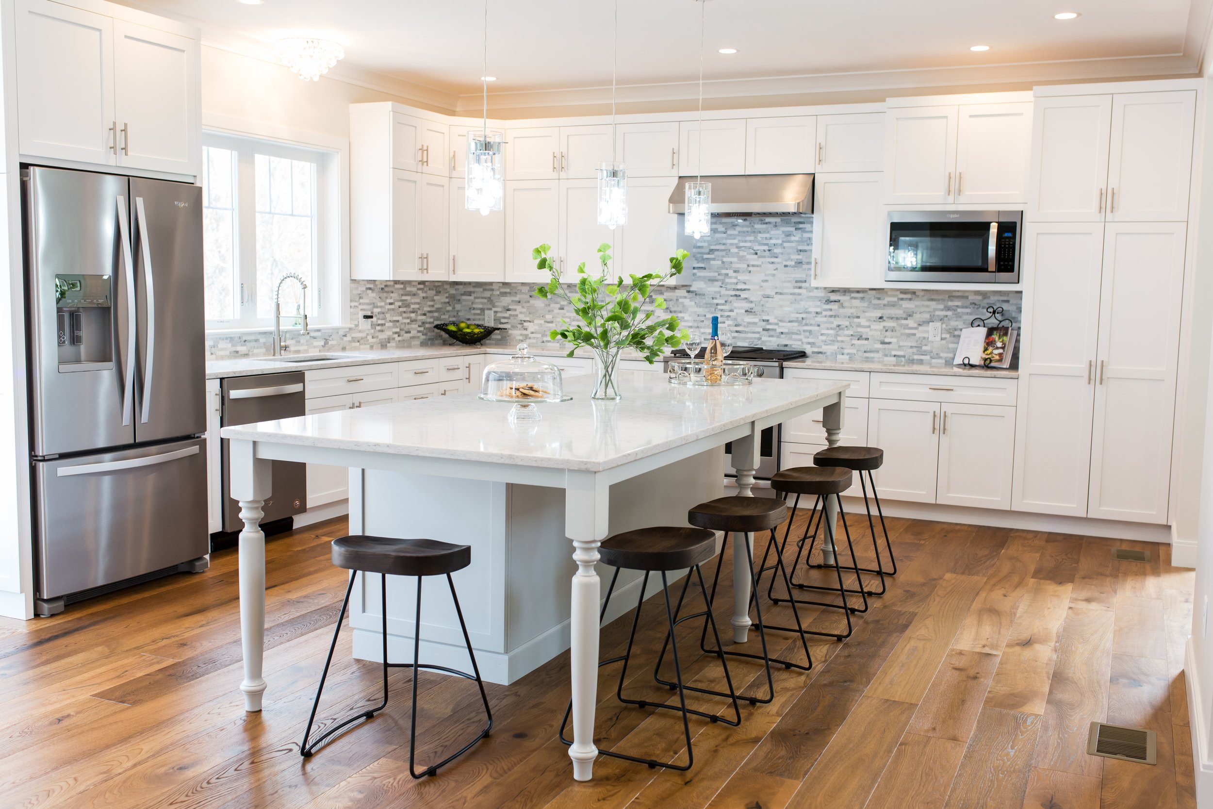 Modern kitchen with white cabinets, stainless steel refrigerator, marble island with bar stools, tiled backsplash, and hardwood floor.