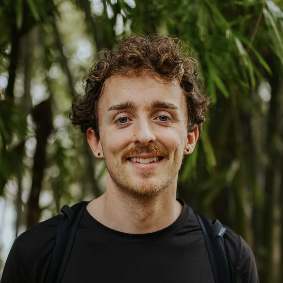 A young man with curly brown hair, mustache, and beard smiling outdoors with green foliage in the background.