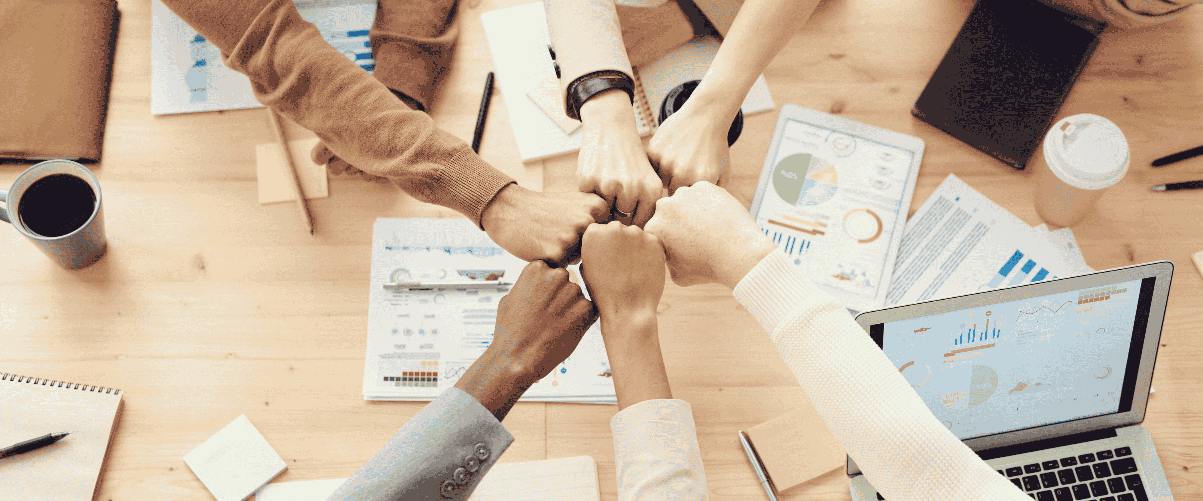 Teamwork with diverse hands fist-bumping over a table with charts, a coffee mug, and a laptop.