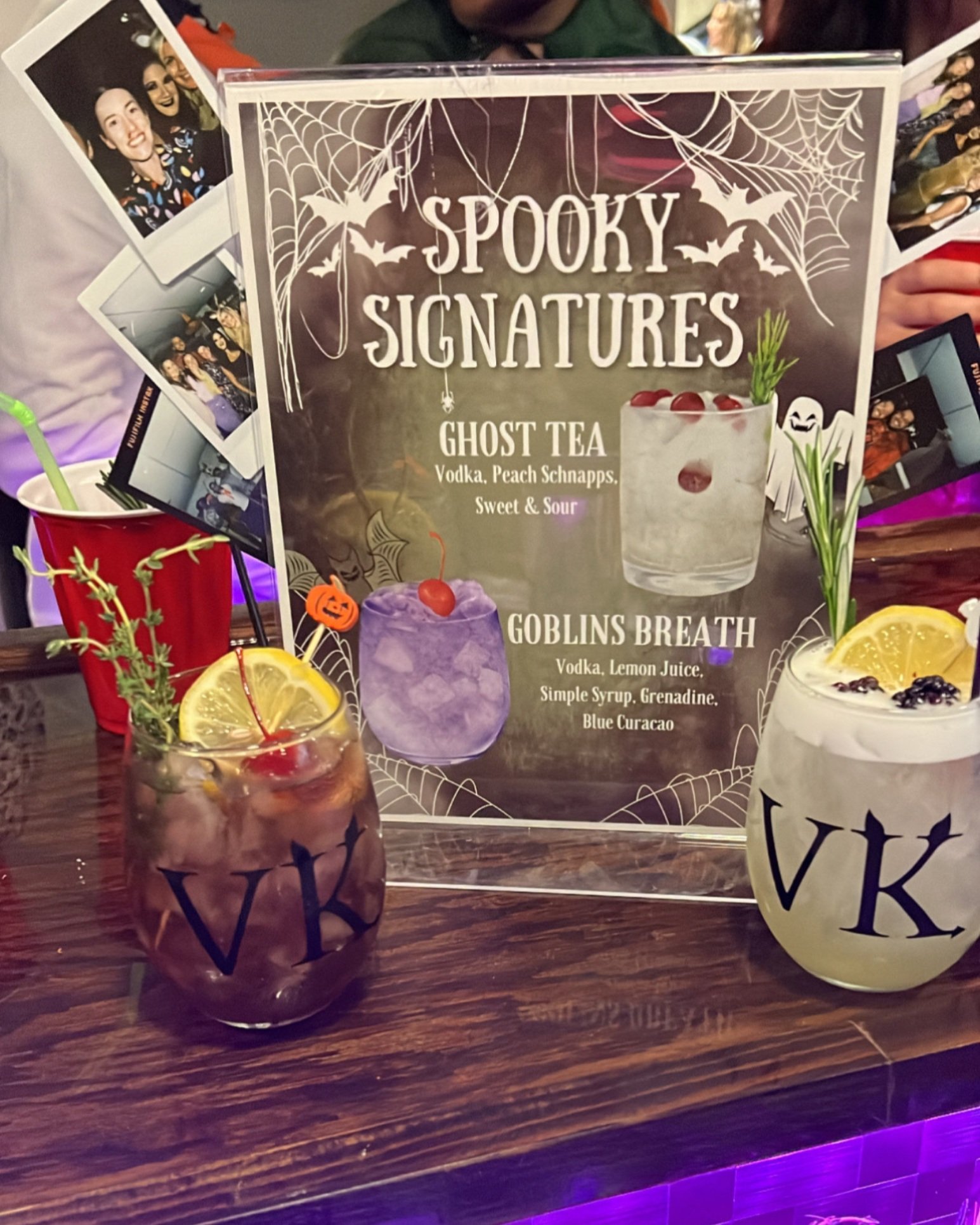 Two Halloween-themed cocktails on a bar table with a sign for spooky signatures, featuring drinks called Ghost Tea and Goblins Breath, surrounded by black and orange decorations and Polaroid photos.