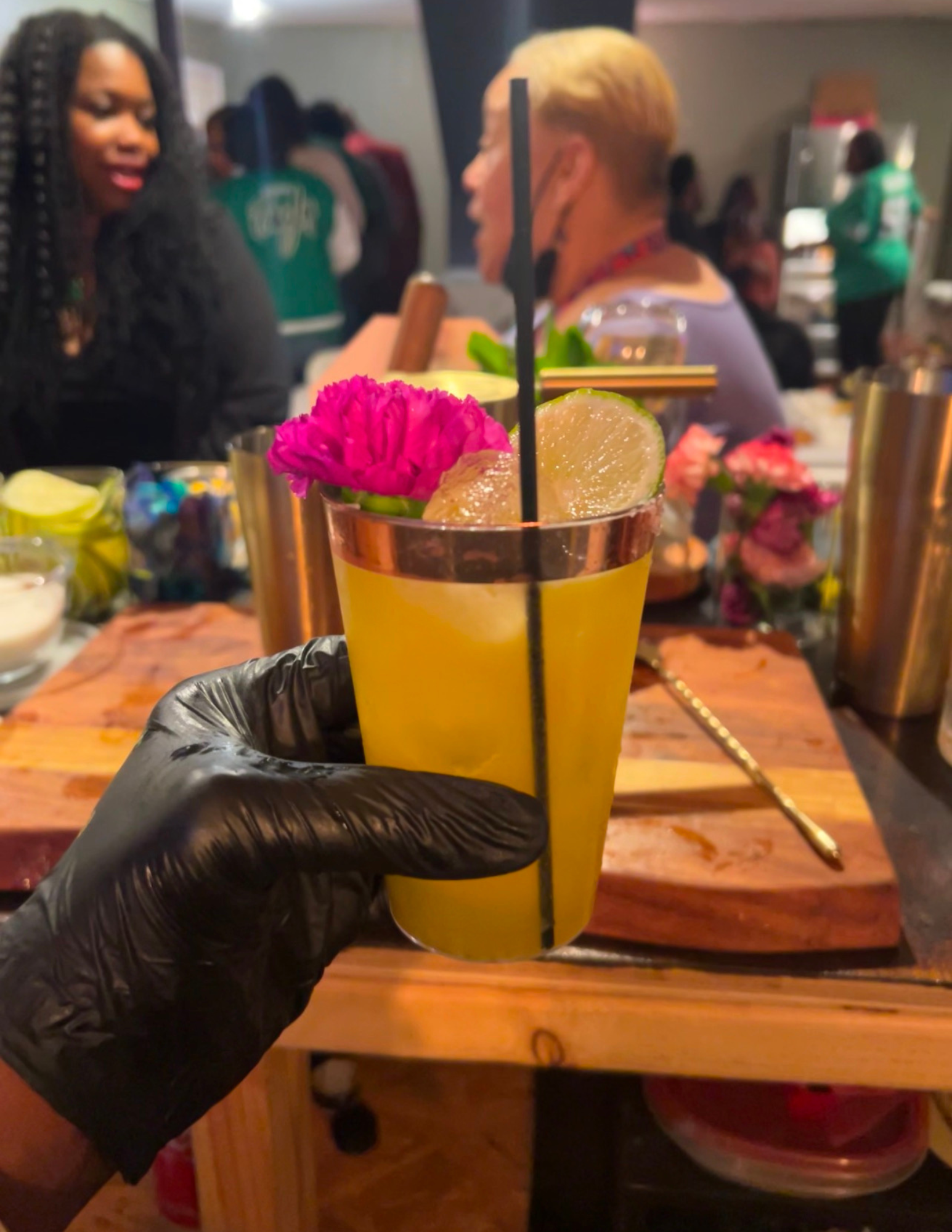 Close-up of a yellow cocktail in a glass garnished with a lime wedge and a pink flower, held by a person wearing a black glove at a restaurant or bar setting with people in the background.
