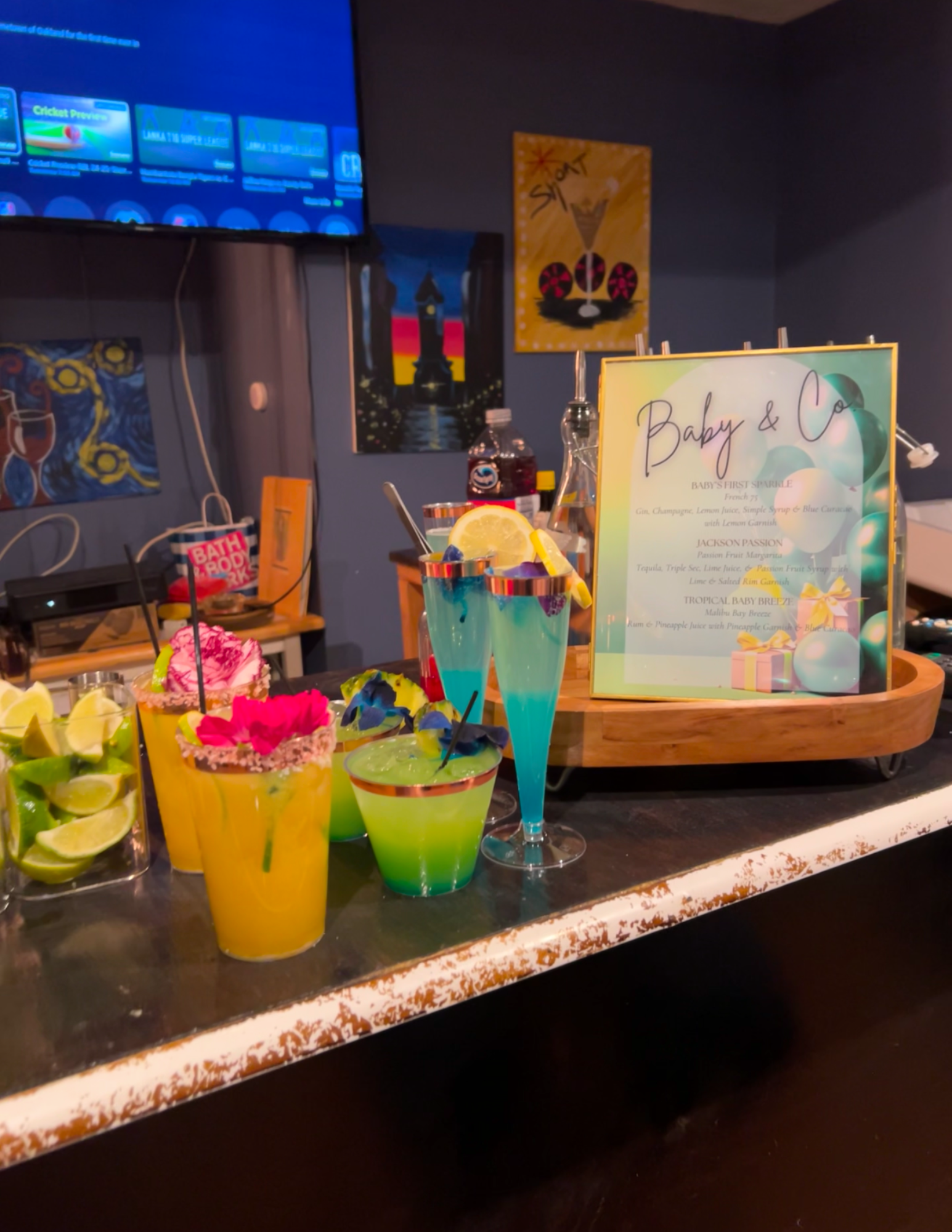Colorful cocktails with lemon garnishes on a bar counter, next to a framed menu and lemon slices, with artwork and a TV screen in the background.