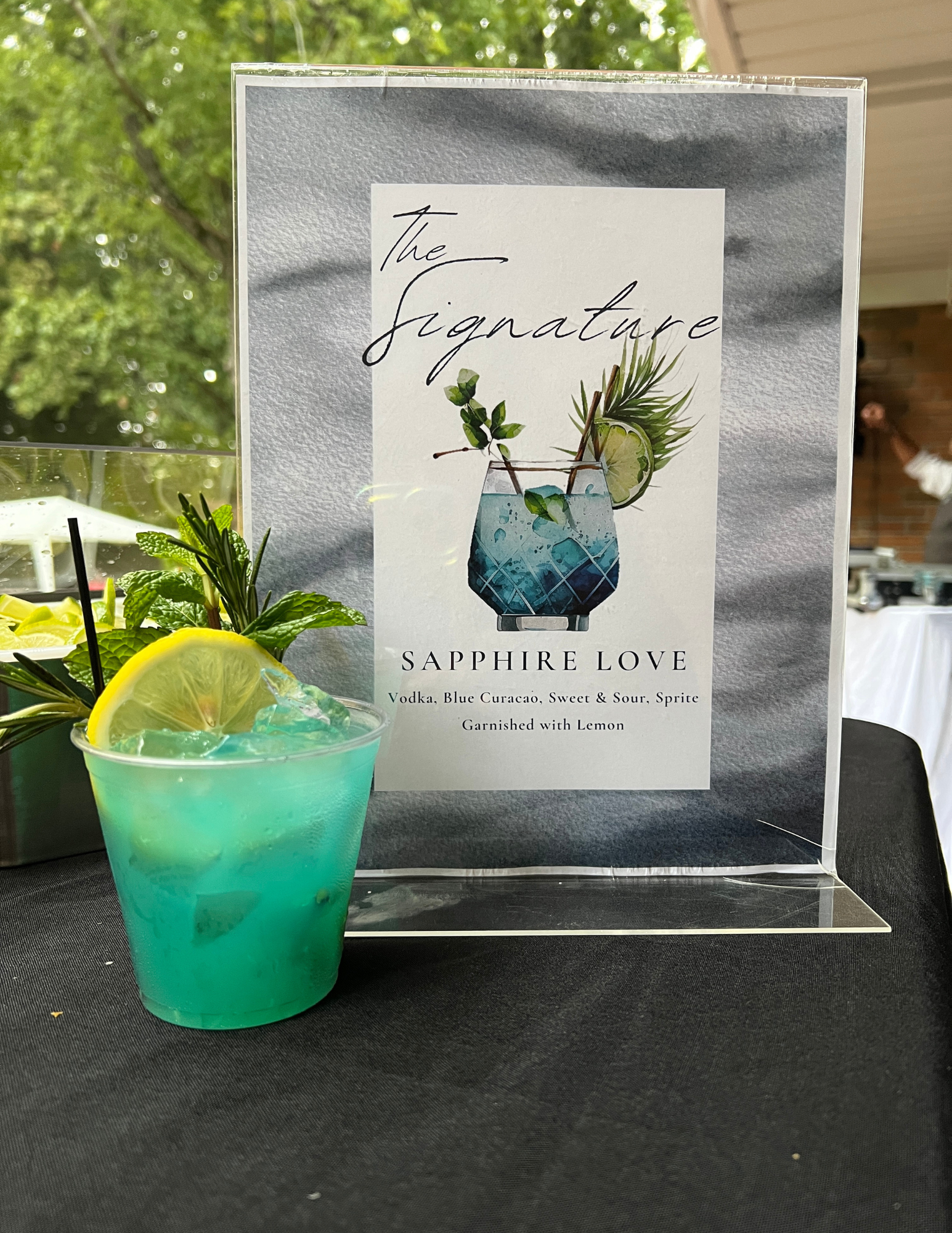 A turquoise cocktail garnished with a lemon slice, mint leaves, and a sprig of rosemary on a black table, with a sign behind it featuring a picture of a cocktail and the words "The Signature Sapphire Love" and ingredients listed.