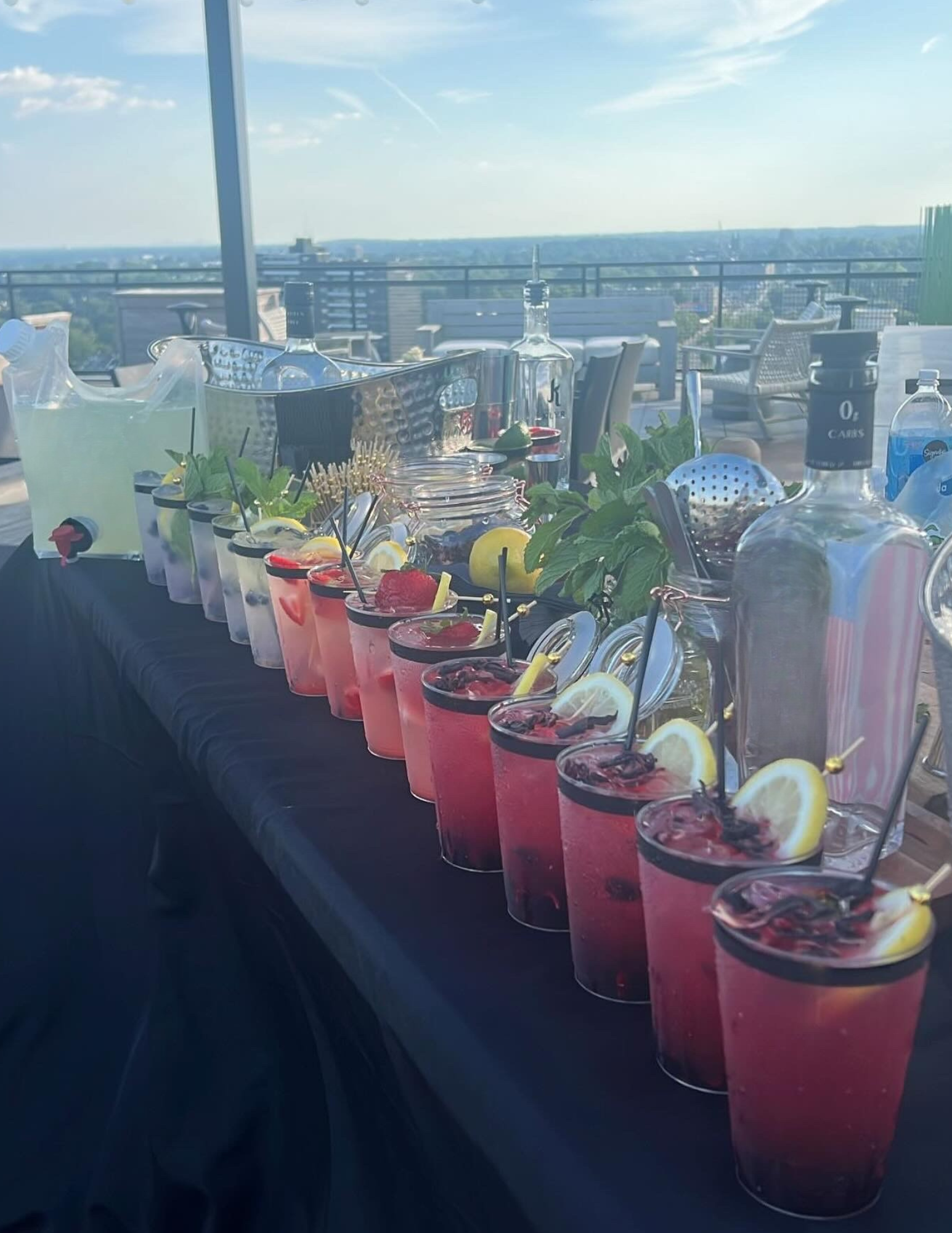 A rooftop table with various colorful cocktails garnished with lemon slices and berries, sets for a gathering with bottles, drink pitchers, and green foliage, overlooking a city skyline and blue sky.