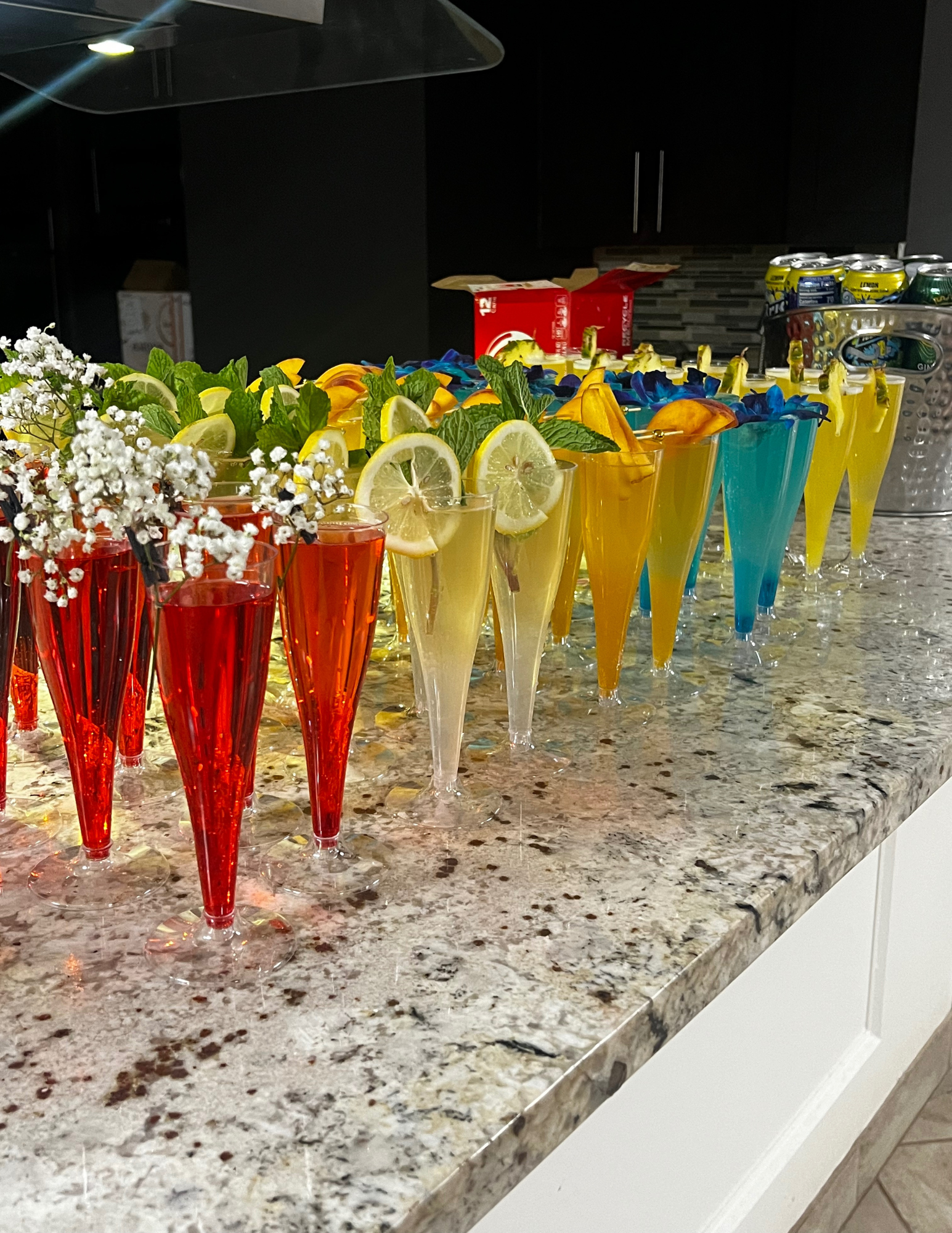 A row of colorful cocktail glasses decorated with lemons, mint leaves, and flowers on a granite countertop.