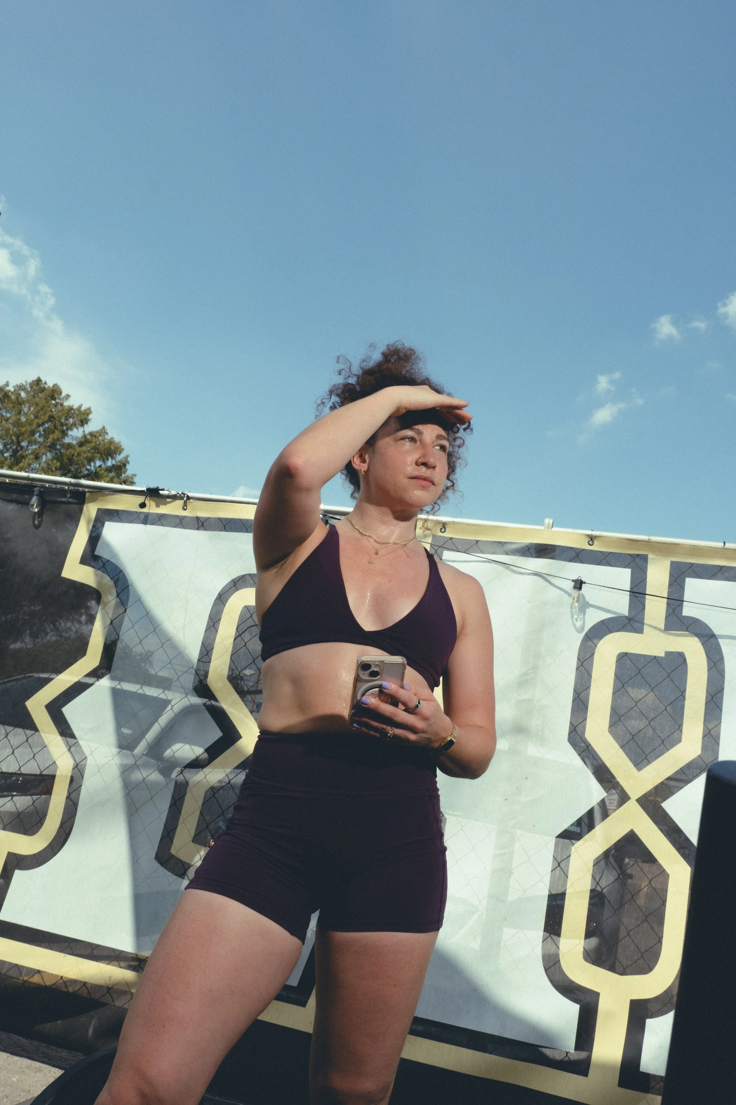 A woman wearing a black sports bra and black shorts, holding a phone, is standing outdoors under a clear blue sky, with a decorative fence or banner and trees in the background.