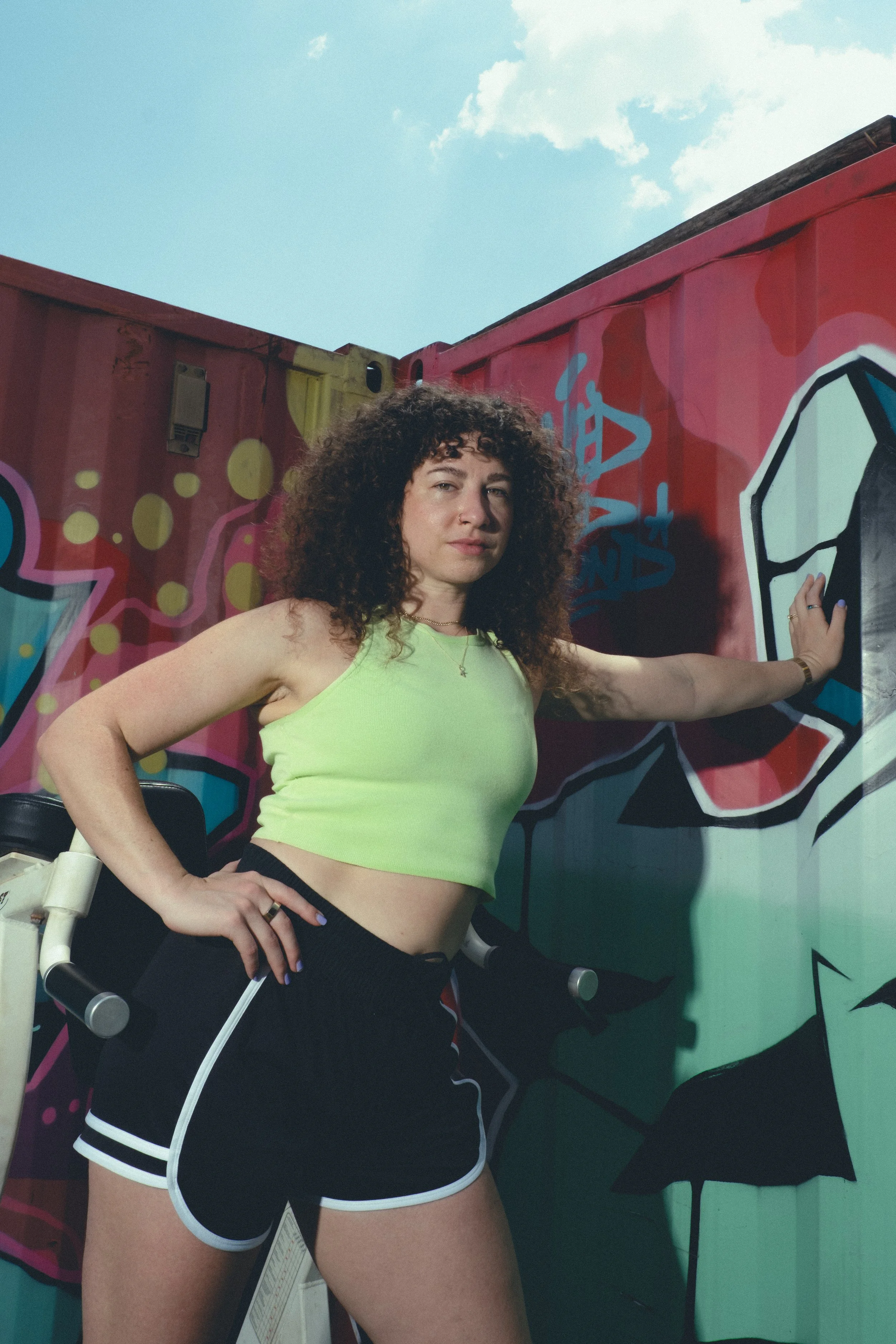A woman with curly hair wearing a yellow crop top and black shorts with white trim, standing in front of a colorful graffiti wall under a blue sky with clouds.