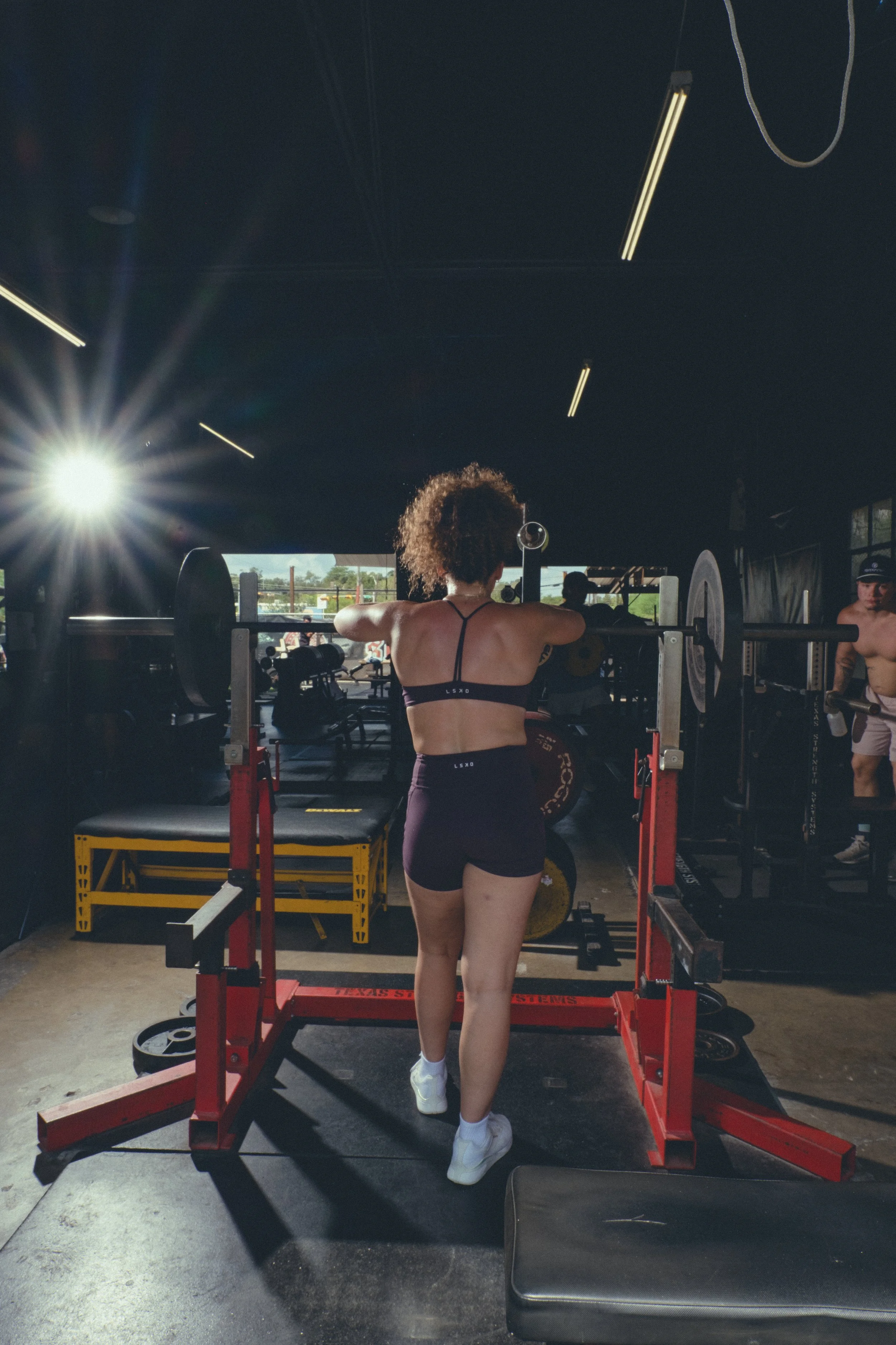 Woman with curly hair lifting weights in a gym, facing away, wearing a sports bra and shorts. Gym equipment and other people are in the background.