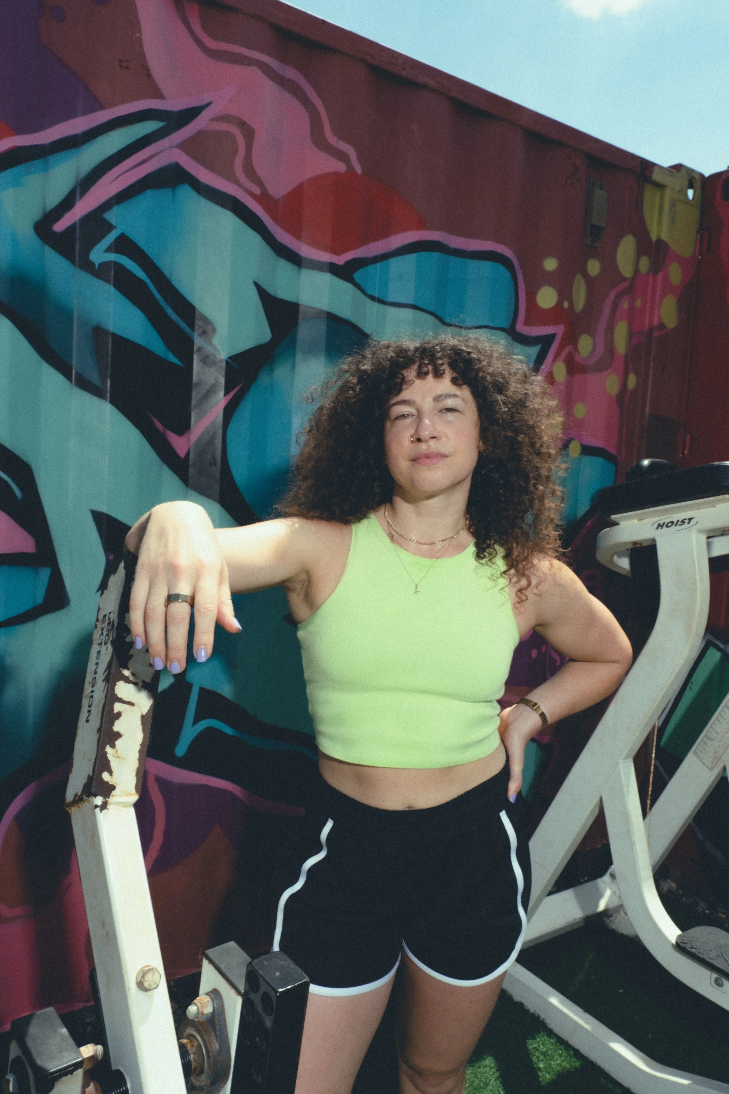 A woman with curly hair wearing a neon green sports bra and black shorts with white trim at the gym, standing in front of a colorful graffiti mural, holding a dumbbell.