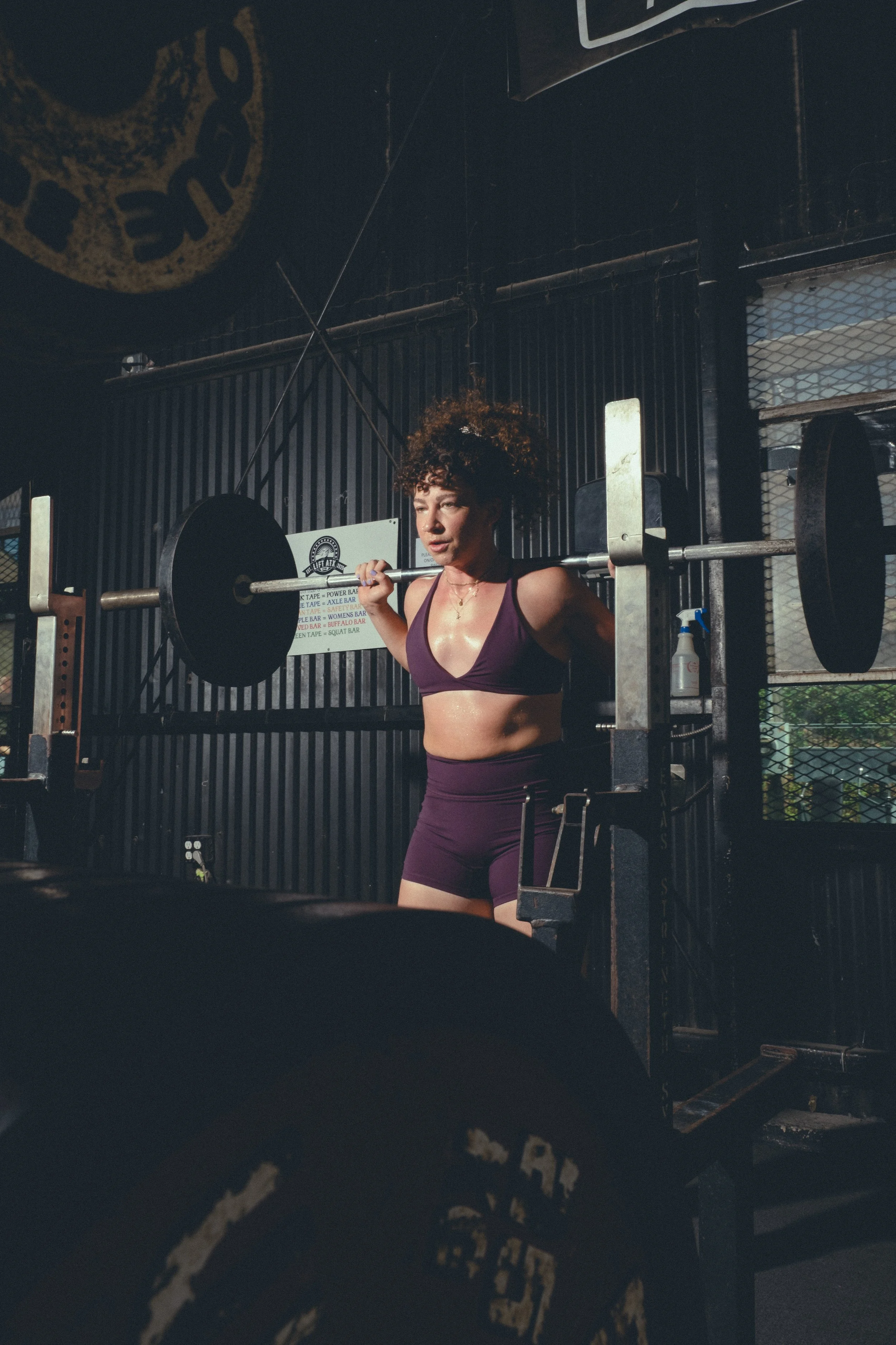 A woman with curly hair and light skin is lifting a barbell during her workout in a gym. She is wearing a purple sports bra and purple shorts, and the gym has black walls with graffiti-style writing and a large logo in the background.