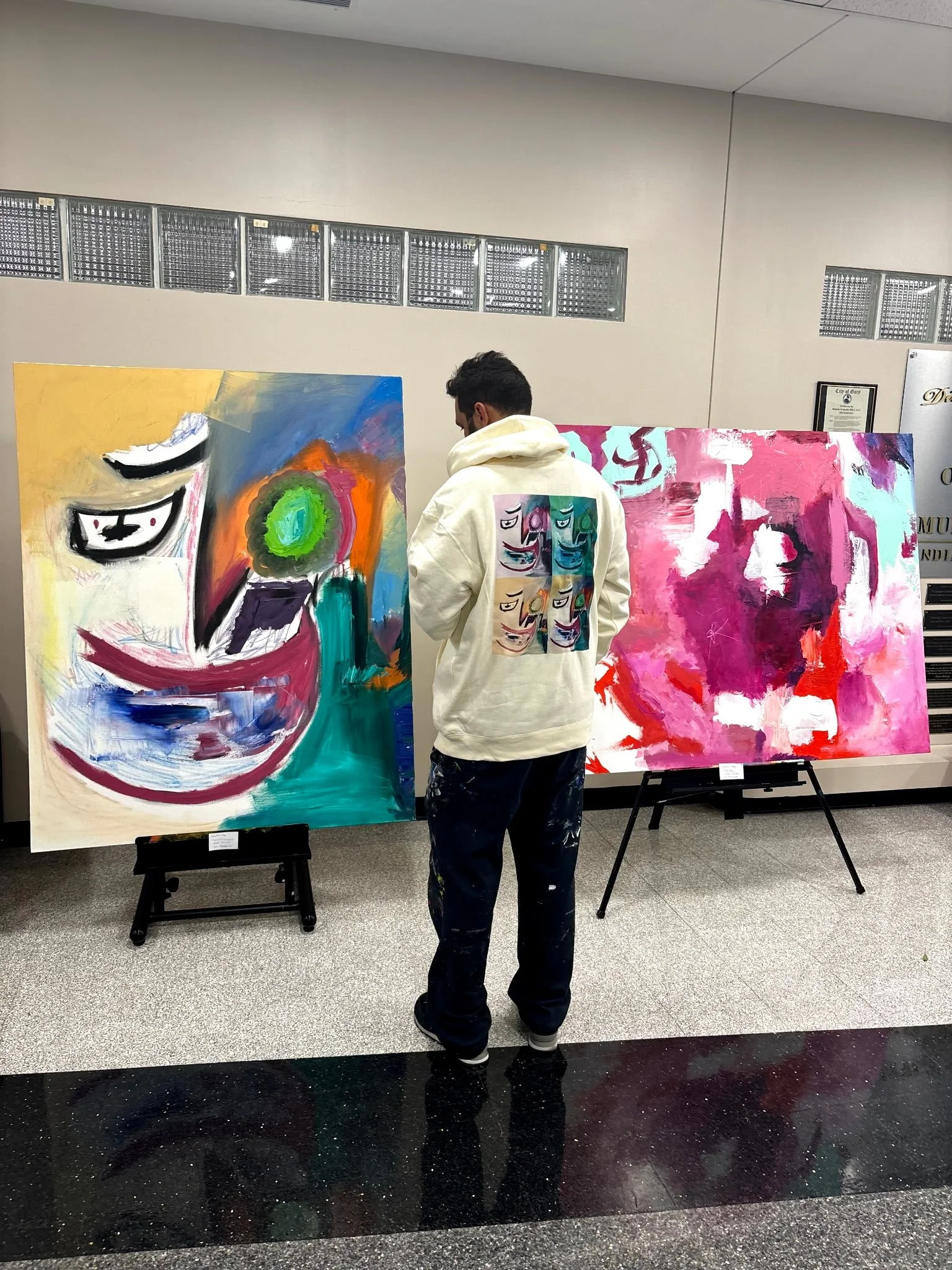 An artist in a white hoodie with six colorful, abstract face designs on the back stands in front of two large abstract paintings displayed on easels in an indoor art gallery. The paintings feature bold colors and expressive brushstrokes.