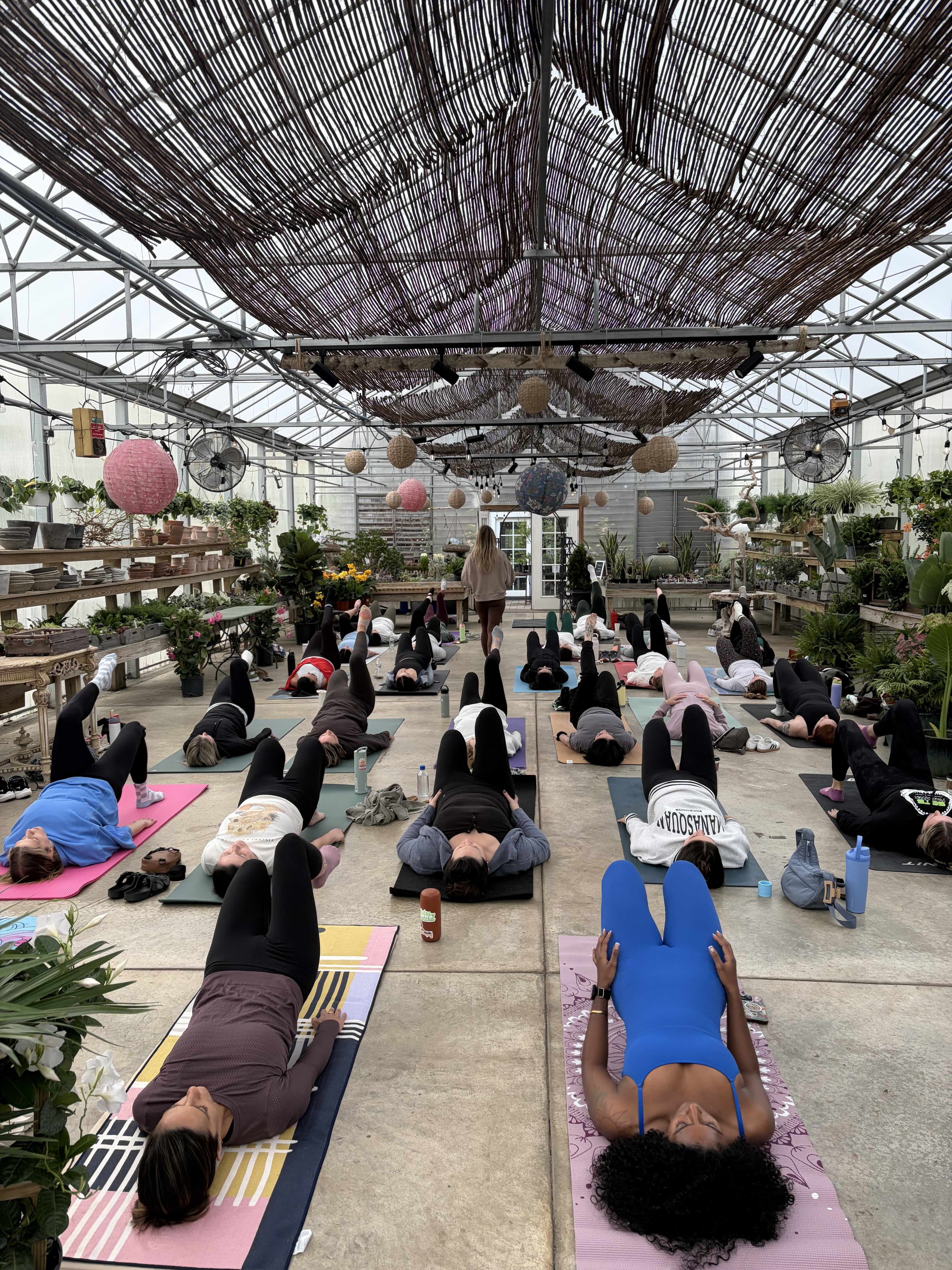 Group of people participating in a yoga class inside a greenhouse, lying on their backs on yoga mats with legs raised, with some facing upward and others facing downward.