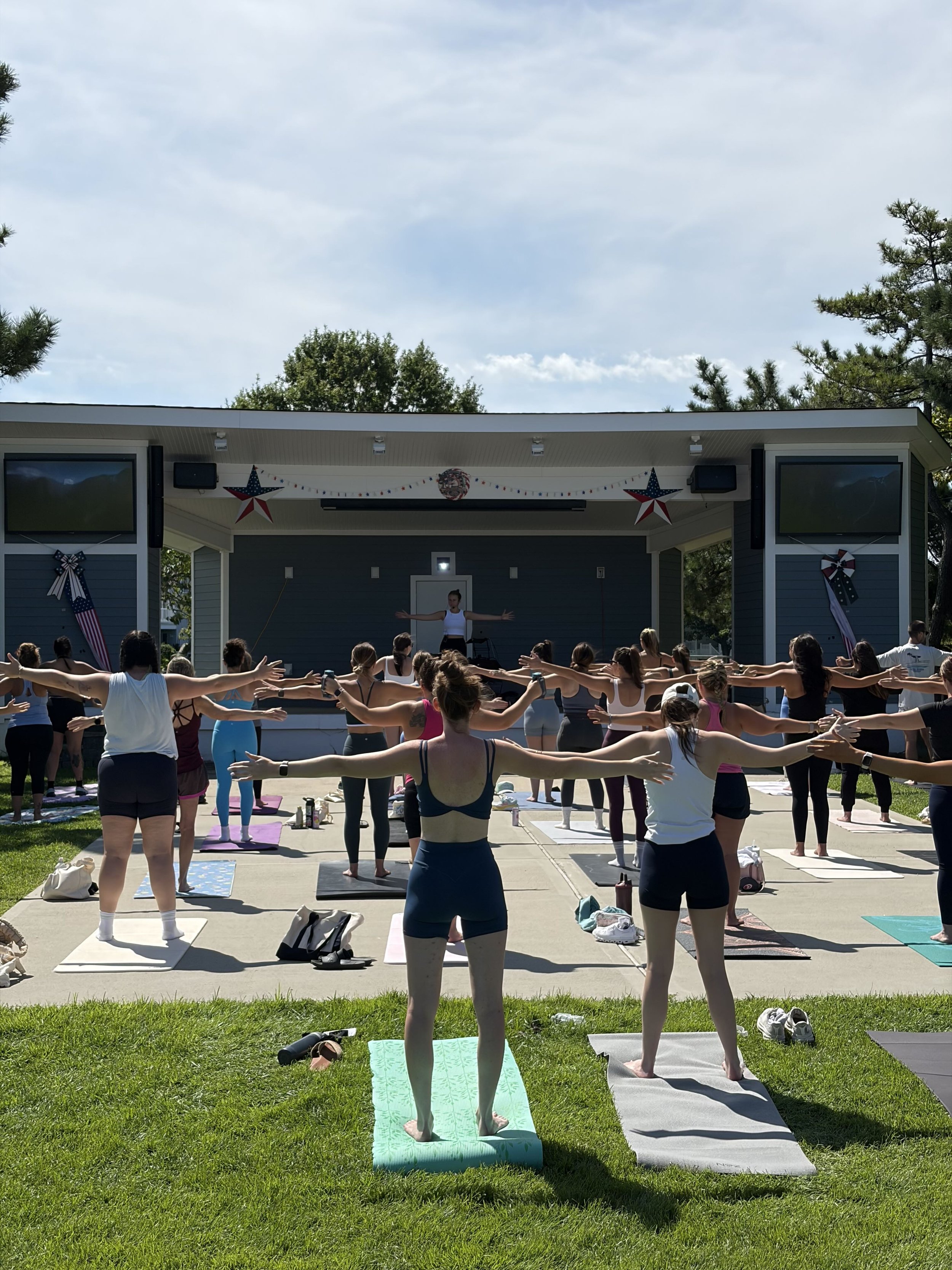 A group of people participating in an outdoor yoga class on a sunny day. They are standing on yoga mats, doing a pose with their arms extended and facing a man leading the class on a stage decorated with patriotic themed hanging stars and banners.