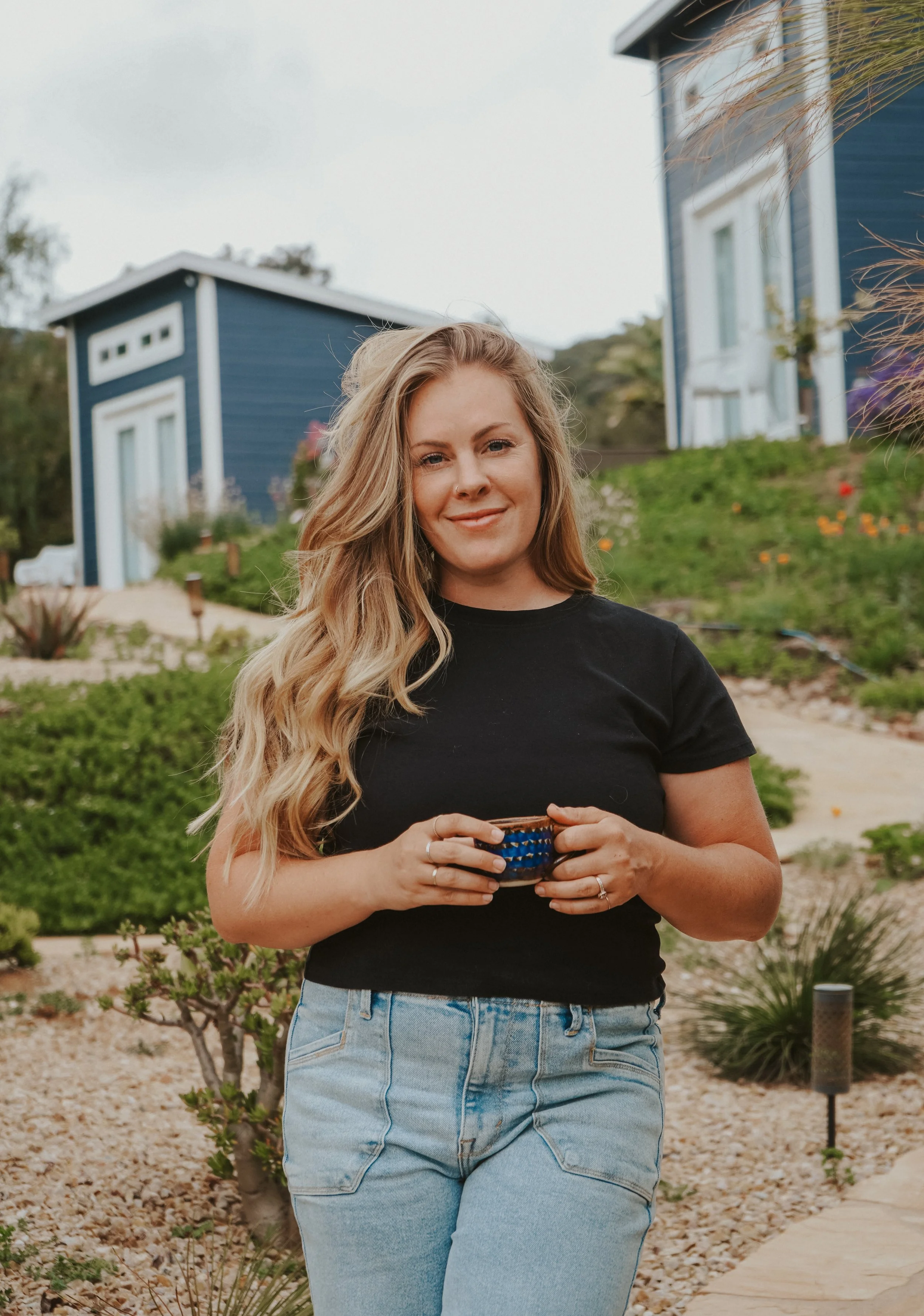 A woman with long blonde hair holding a small coffee cup, standing outdoors in front of a blue house with a landscaped garden.