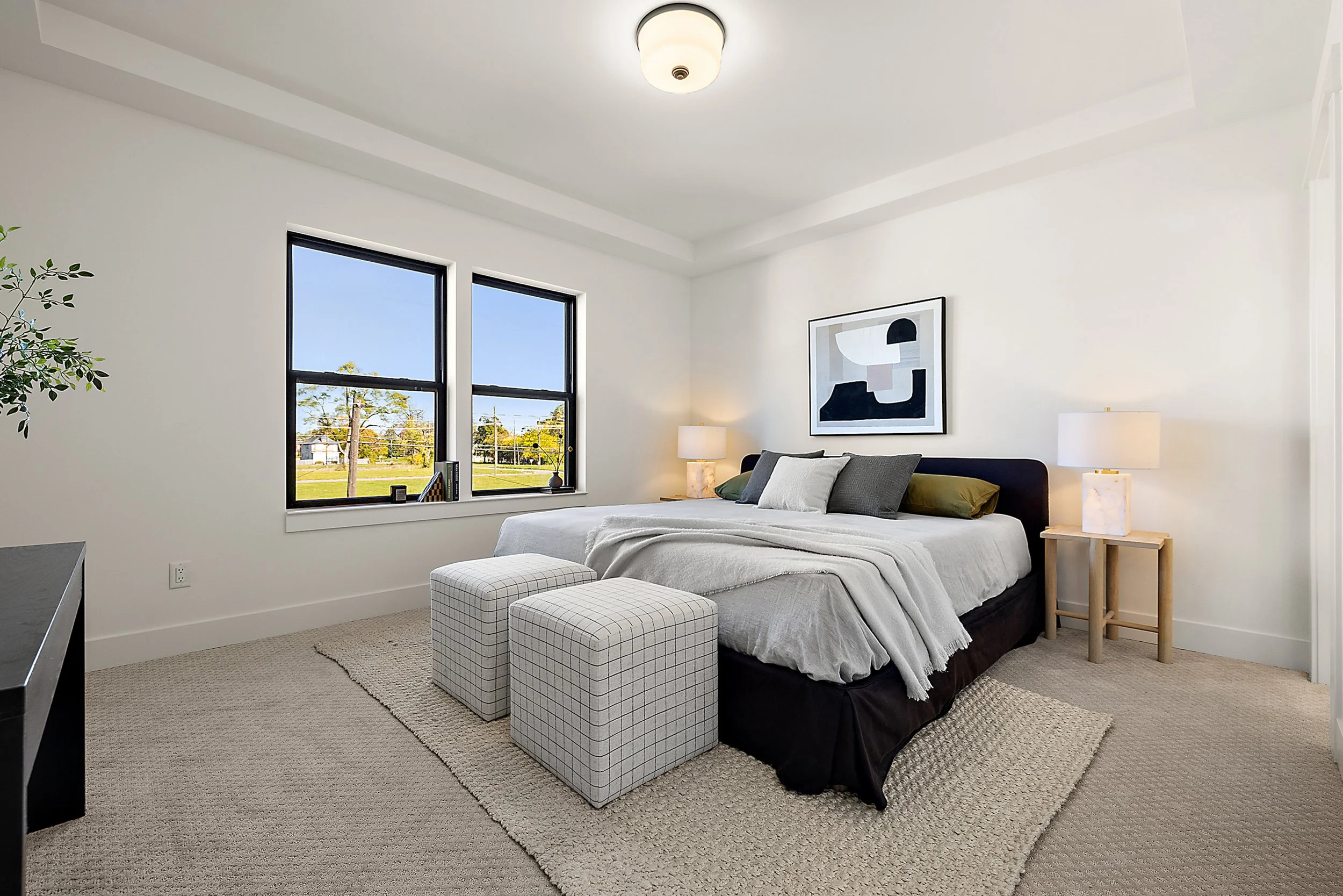 A modern bedroom with a large bed, two checkered ottomans, bedside lamps, a framed abstract art piece, a window showing a sunny outdoor scene, and a beige textured rug.