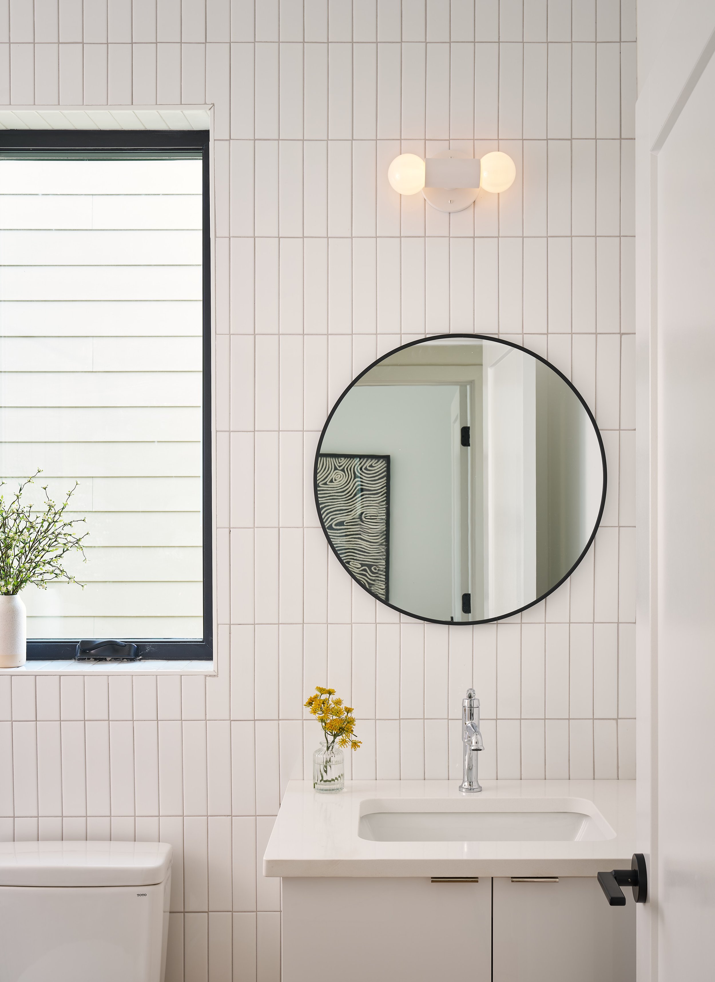 A bright bathroom with white tiled walls, a window with a plant on the windowsill, a round mirror above a white sink, and a light fixture with two globes.