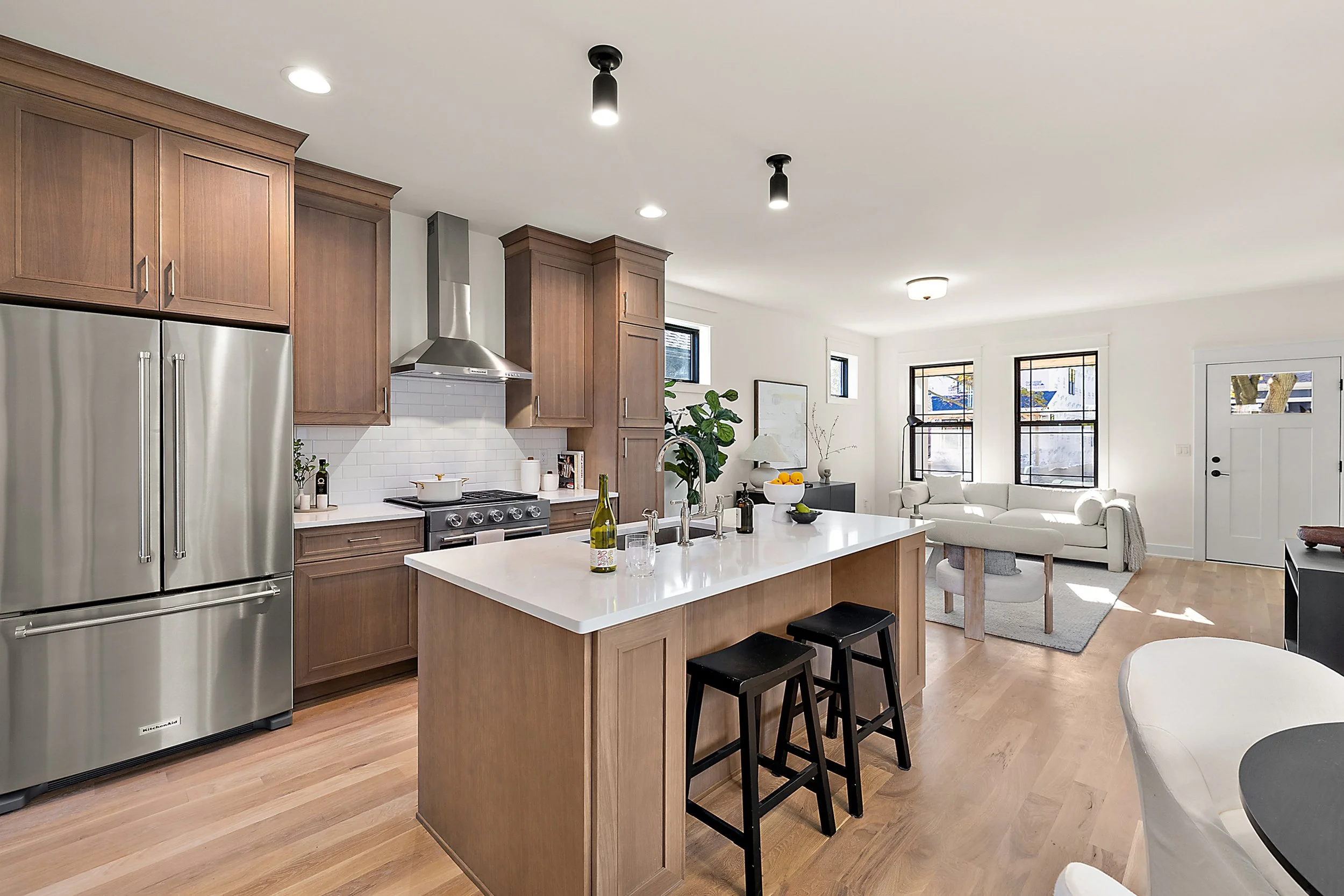 Open-concept kitchen and living room with wooden cabinetry, stainless steel appliances, white island with bar stools, and white sofa, with hardwood floors and black-framed windows.