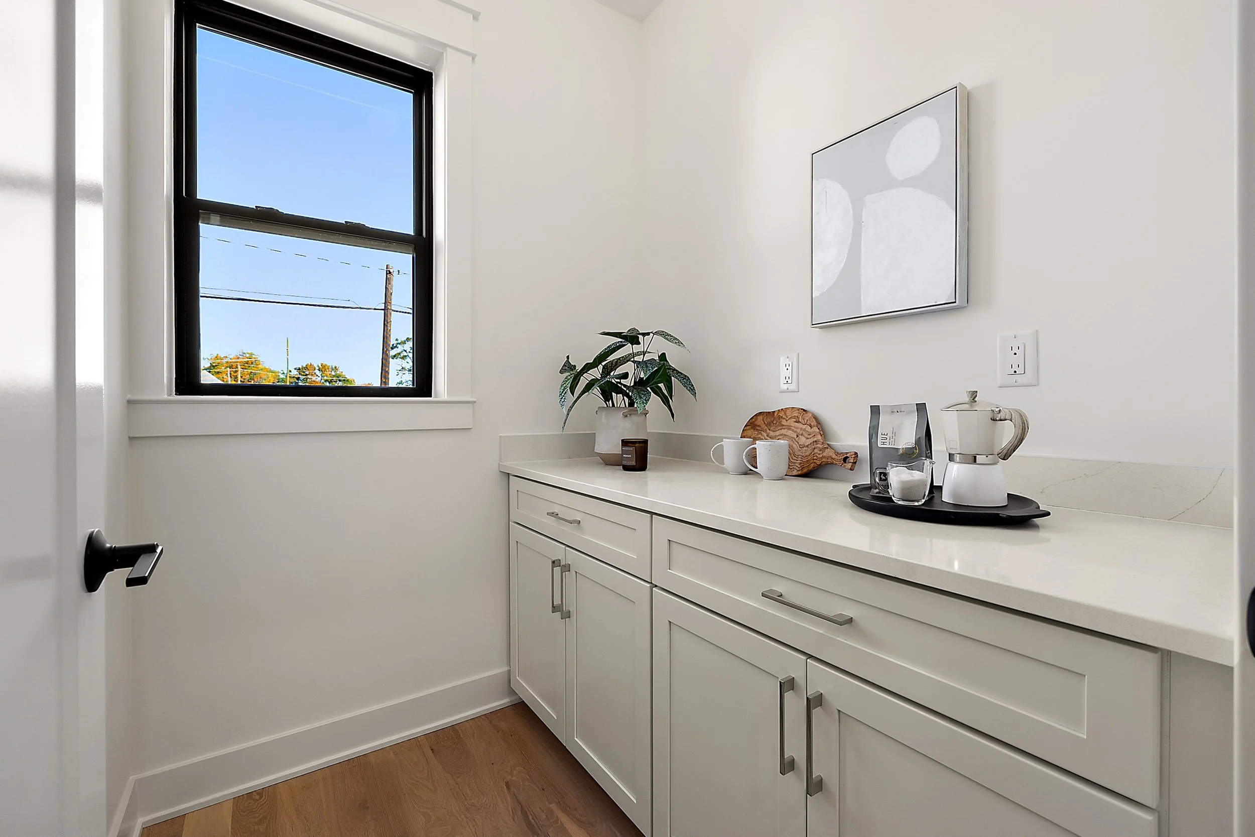 A white kitchen counter with a potted plant, coffee maker, and cups, under a window with a blue sky outside. A piece of abstract wall art hangs on the white wall.