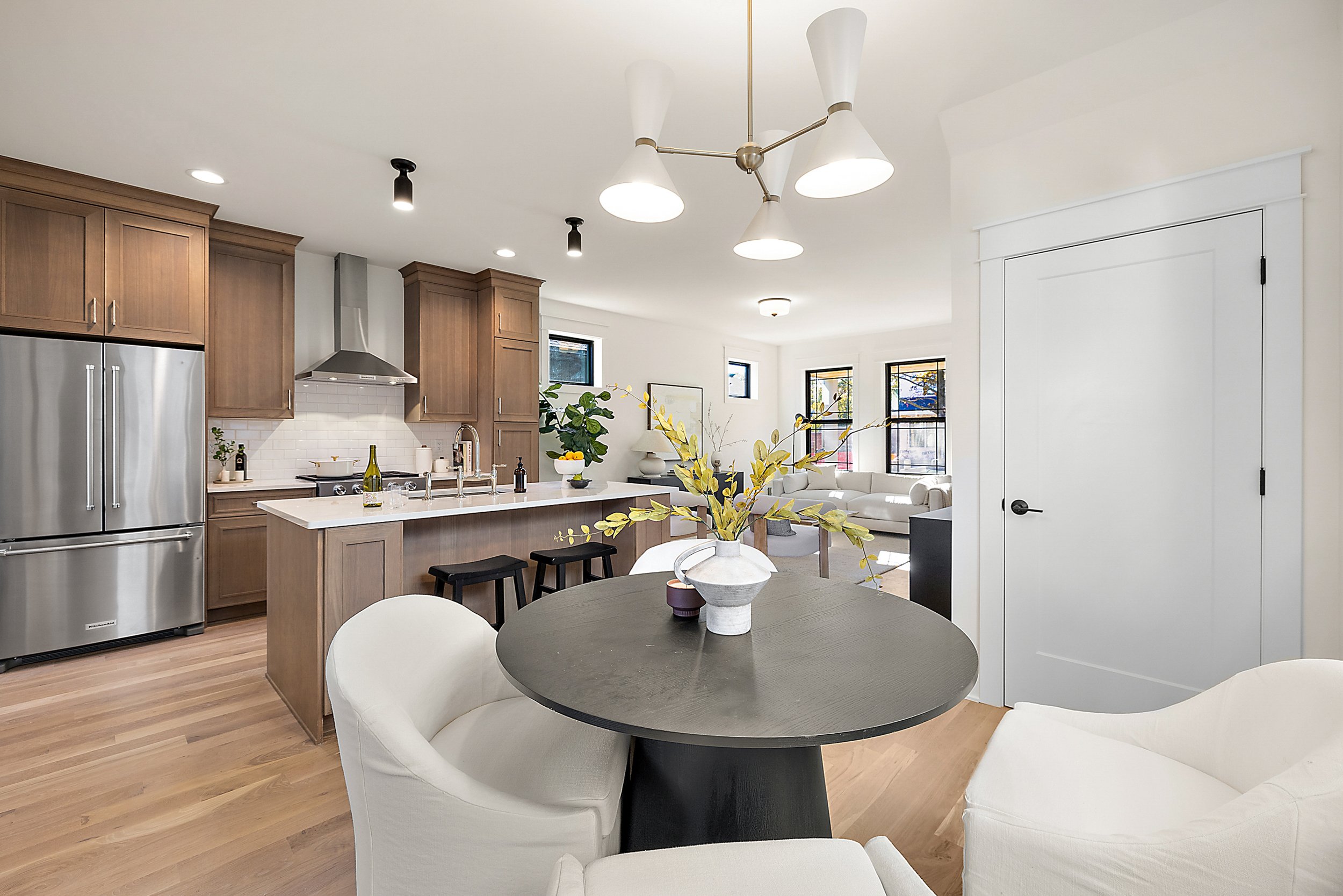 Open-concept kitchen and living room with wooden cabinets, stainless steel refrigerator, island with bar stools, round dining table with white upholstered chairs, and large windows.