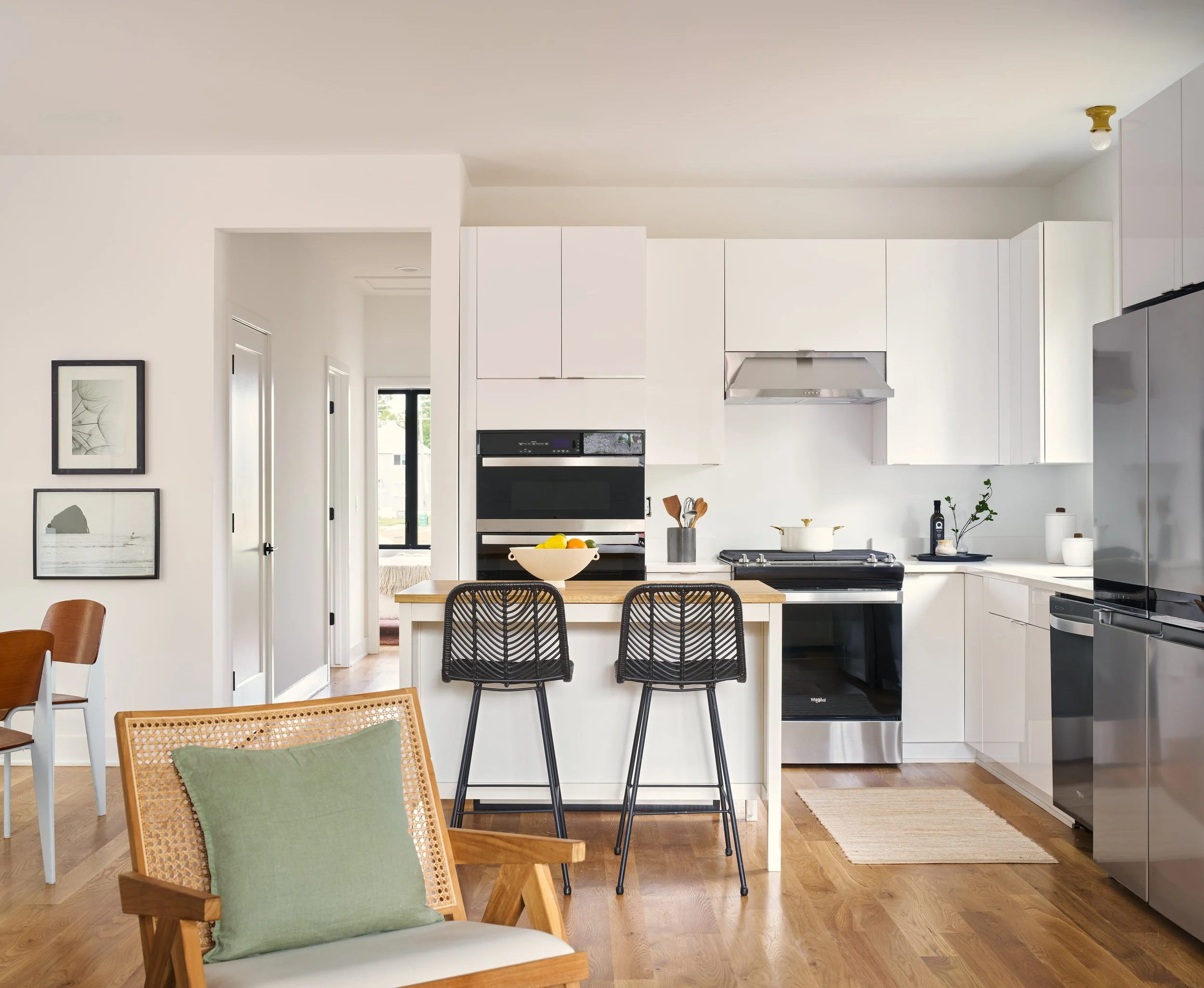 Modern kitchen with white cabinets, stainless steel appliances, black bar stools, wooden floor, and minimalist decor.