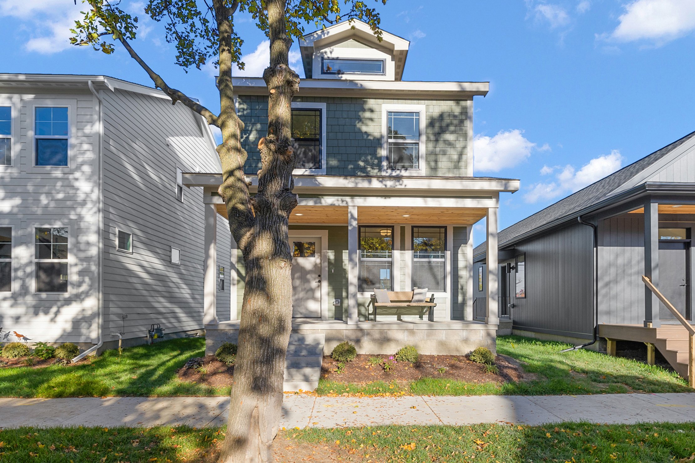 Modern multi-story house with a porch, sitting area, and a large tree in the front yard on a sunny day.