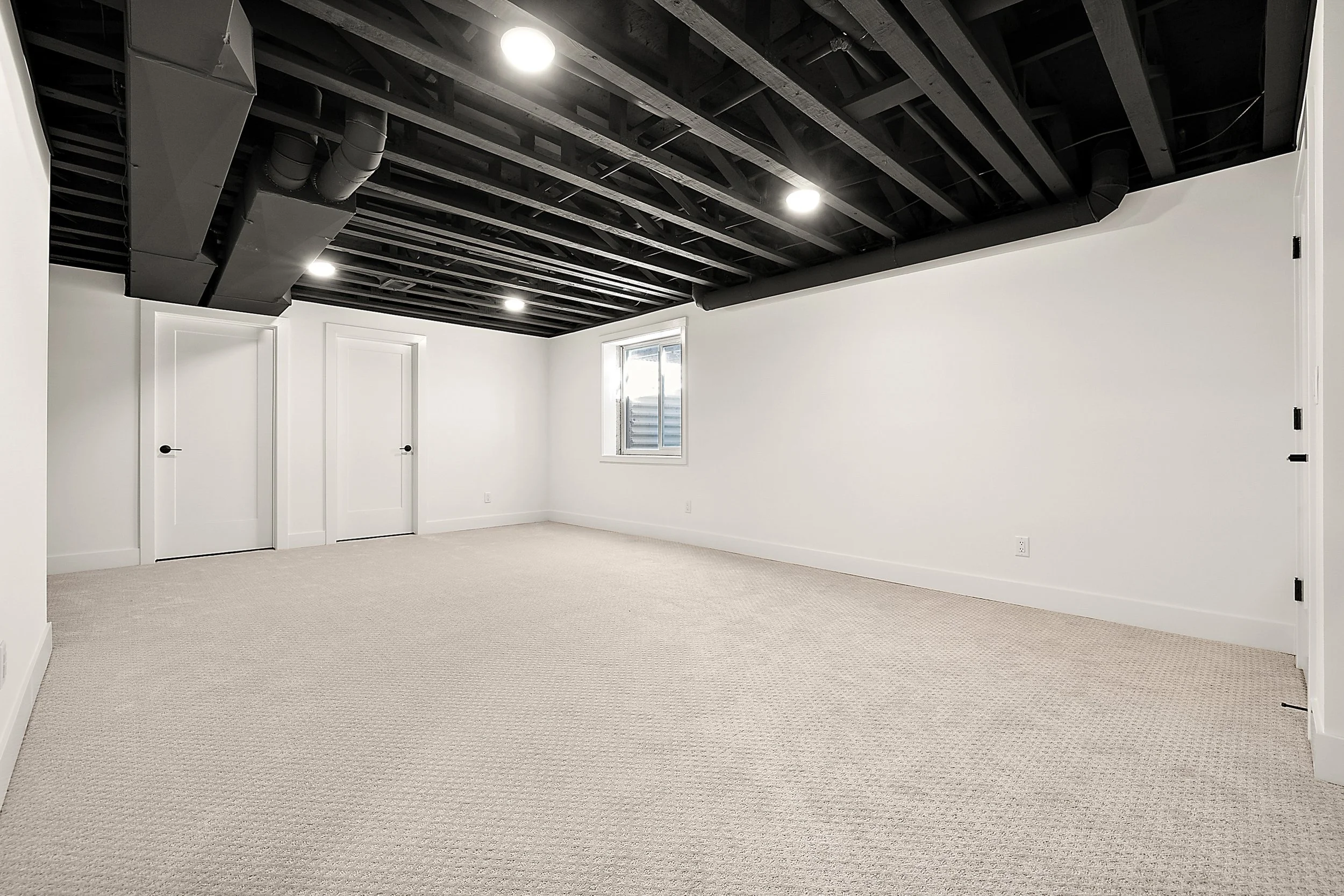 Empty room with white walls, light beige carpet, black ceiling with exposed beams and ductwork, window on one wall, and two closed doors.