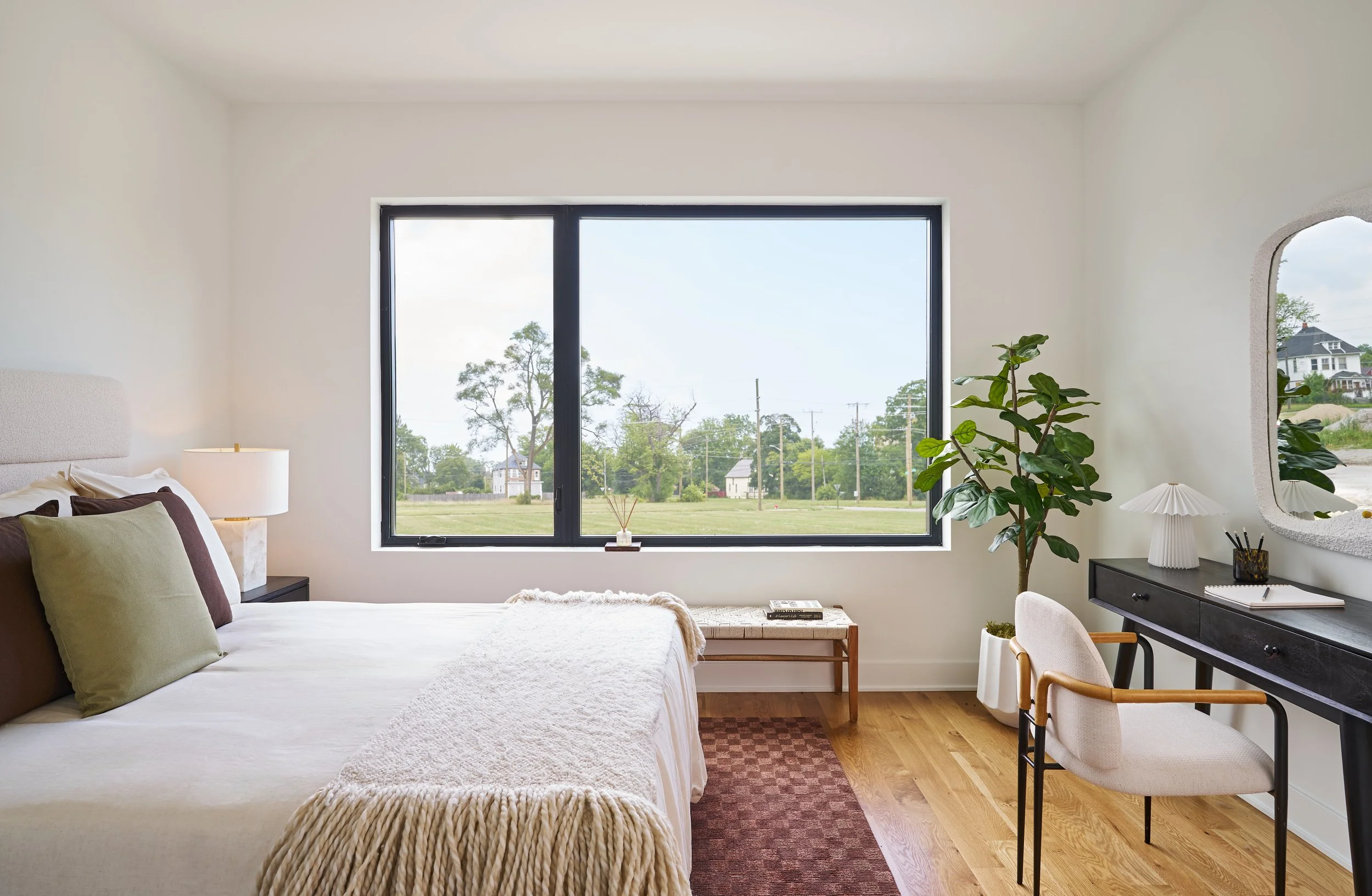Bright bedroom with a large window overlooking a yard with trees and houses, featuring a bed with pillows, a side table with a lamp, a wooden bench with magazine, a tall potted plant, a black desk with lamp and mirror, and an office chair.