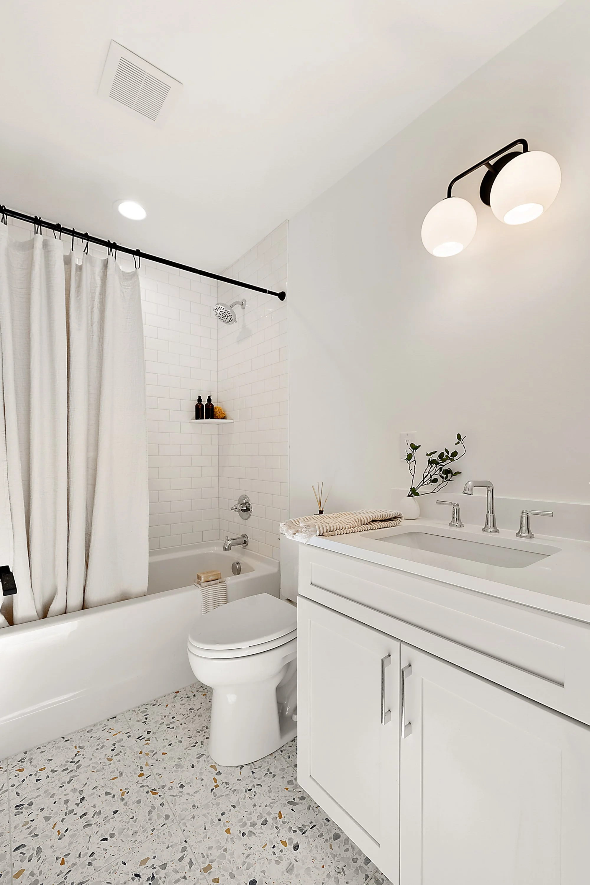 Modern bathroom with white bathtub, shower with white curtain, white toilet, white vanity with a sink, green leafy plant, and contemporary light fixture.