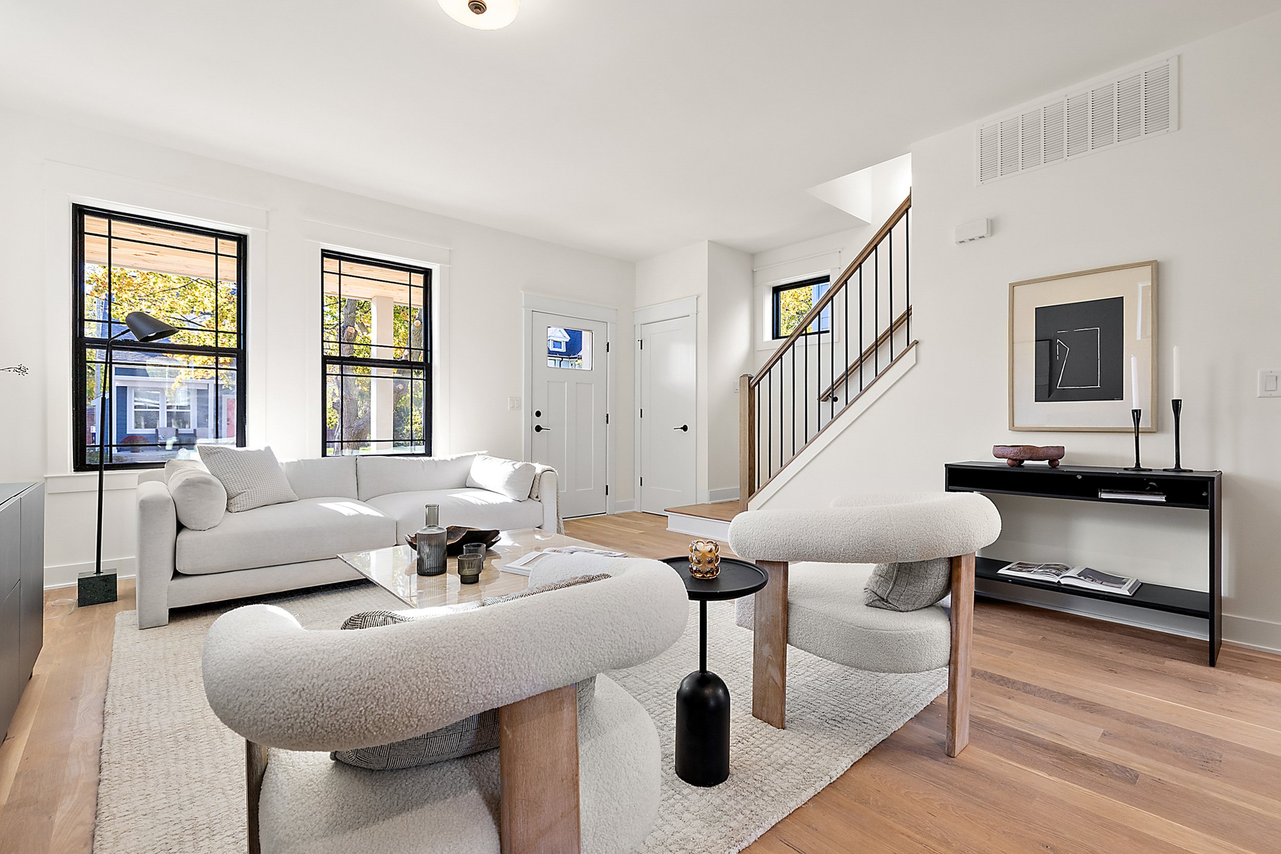 Minimalist living room with white sofa, armchairs, black side table, black console, wooden flooring, large windows, and staircase in background