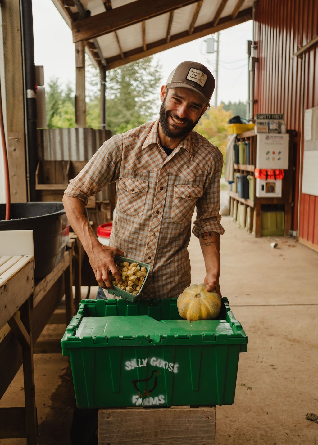 A smiling man wearing a plaid shirt and a cap is holding a container of garlic cloves in one hand and a pumpkin in the other. He is standing in a farm barn with trees visible outside. There is a green box labeled 'Silly Goose Farms' in front of him.