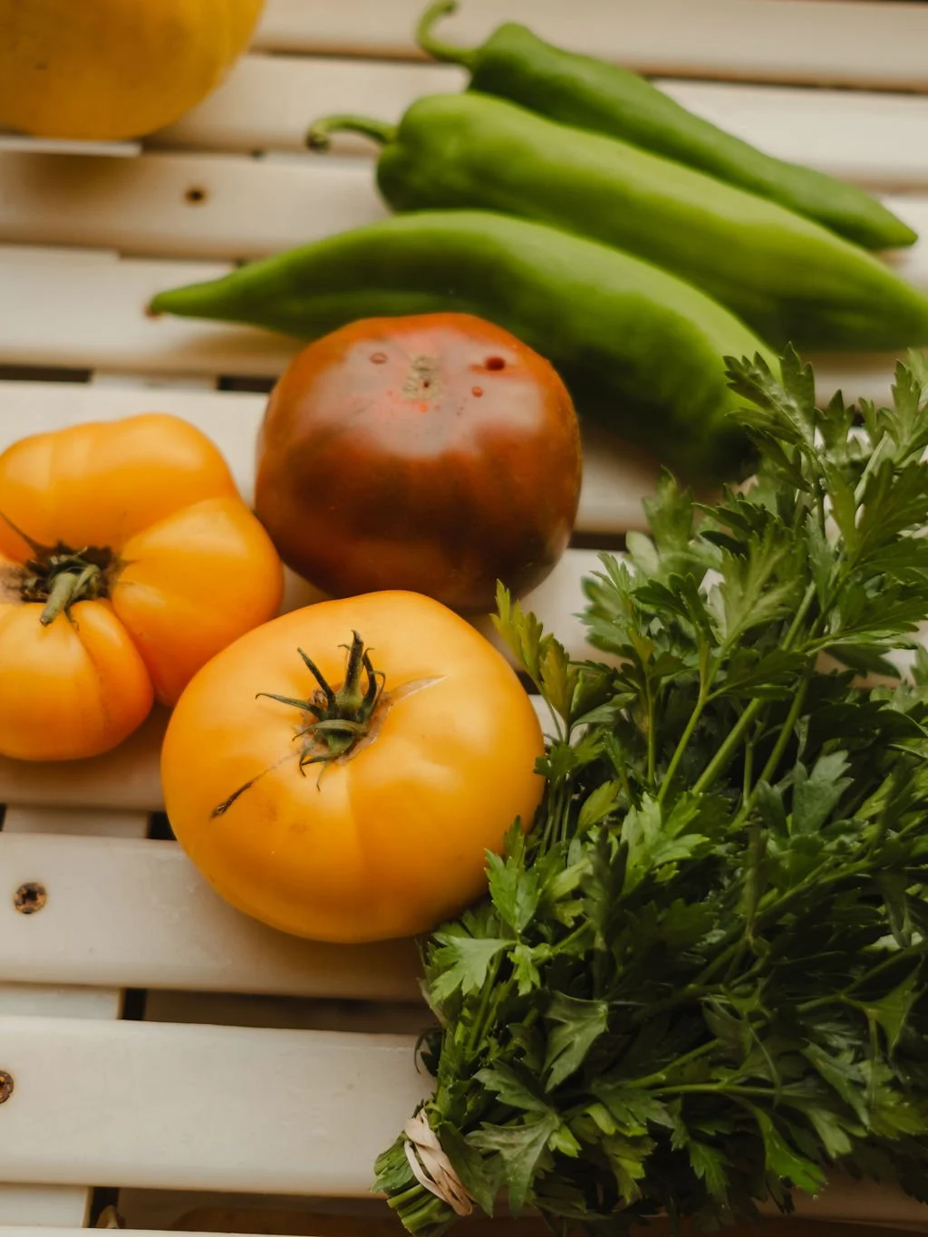 Group of fresh vegetables on a white wooden surface, including yellow tomatoes, a red and black heirloom tomato, green chili peppers, and parsley.