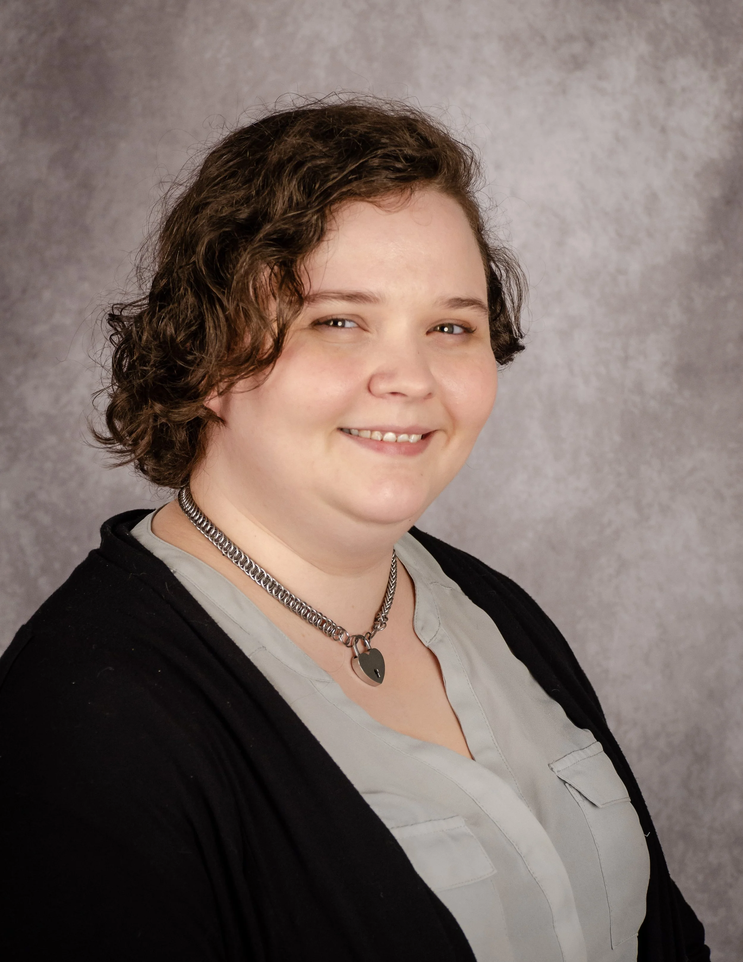 Person with short curly hair wearing a necklace and a light-colored top with a dark cardigan, smiling in front of a gray textured background.