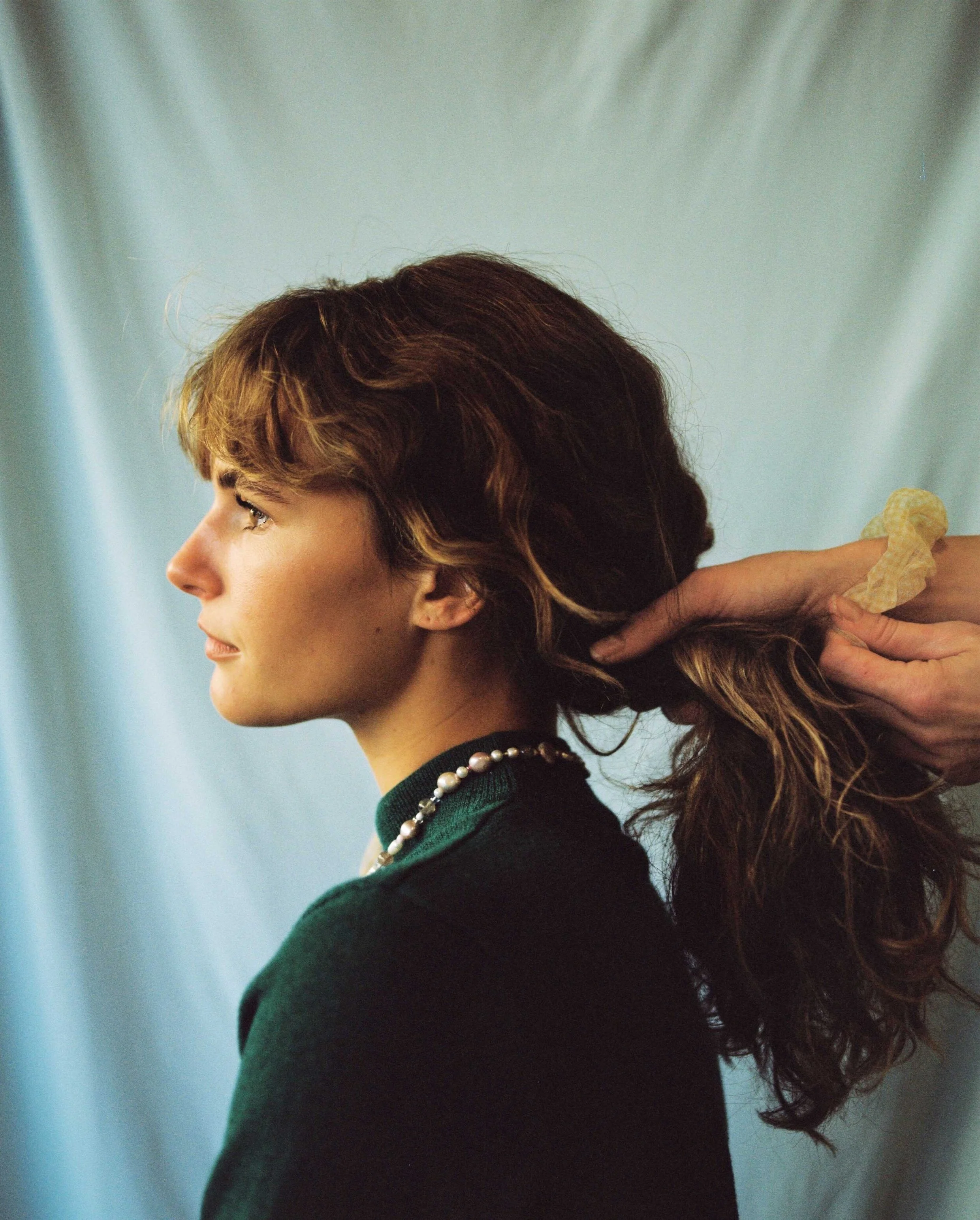 A woman with wavy, shoulder-length brown hair is having her hair styled, with someone holding a section of her hair from the back.