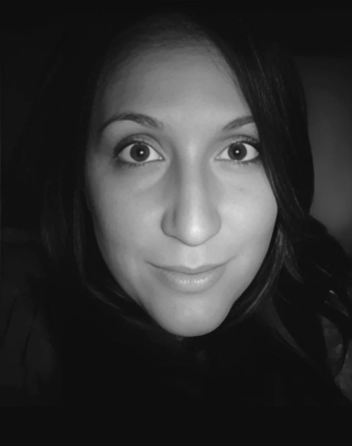 A close-up black and white photo of a woman with light skin, long dark hair, and large eyes, smiling softly.