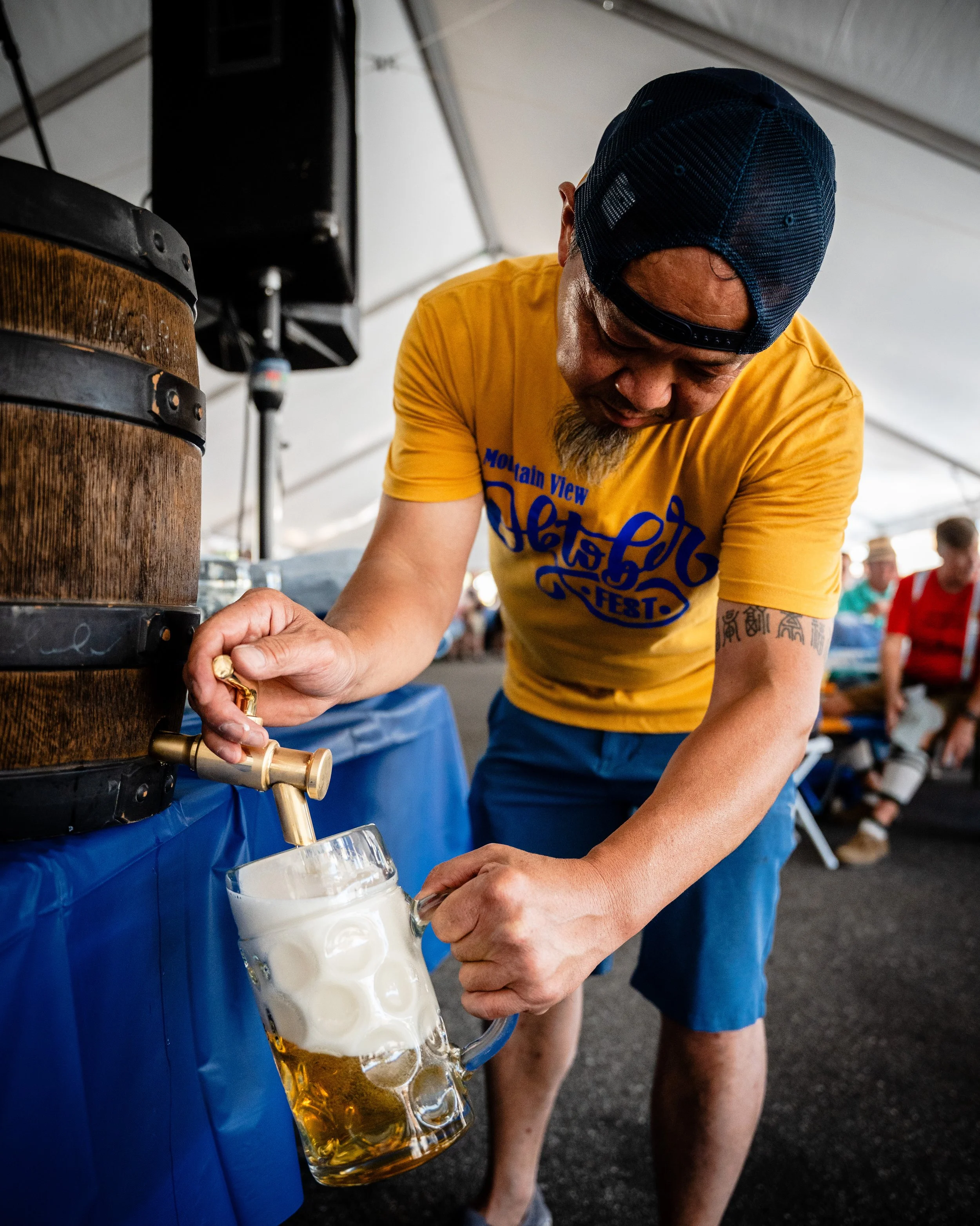 A man in a yellow t-shirt and blue shorts pours beer from a tap into a glass mug at a festival or event under a large tent.