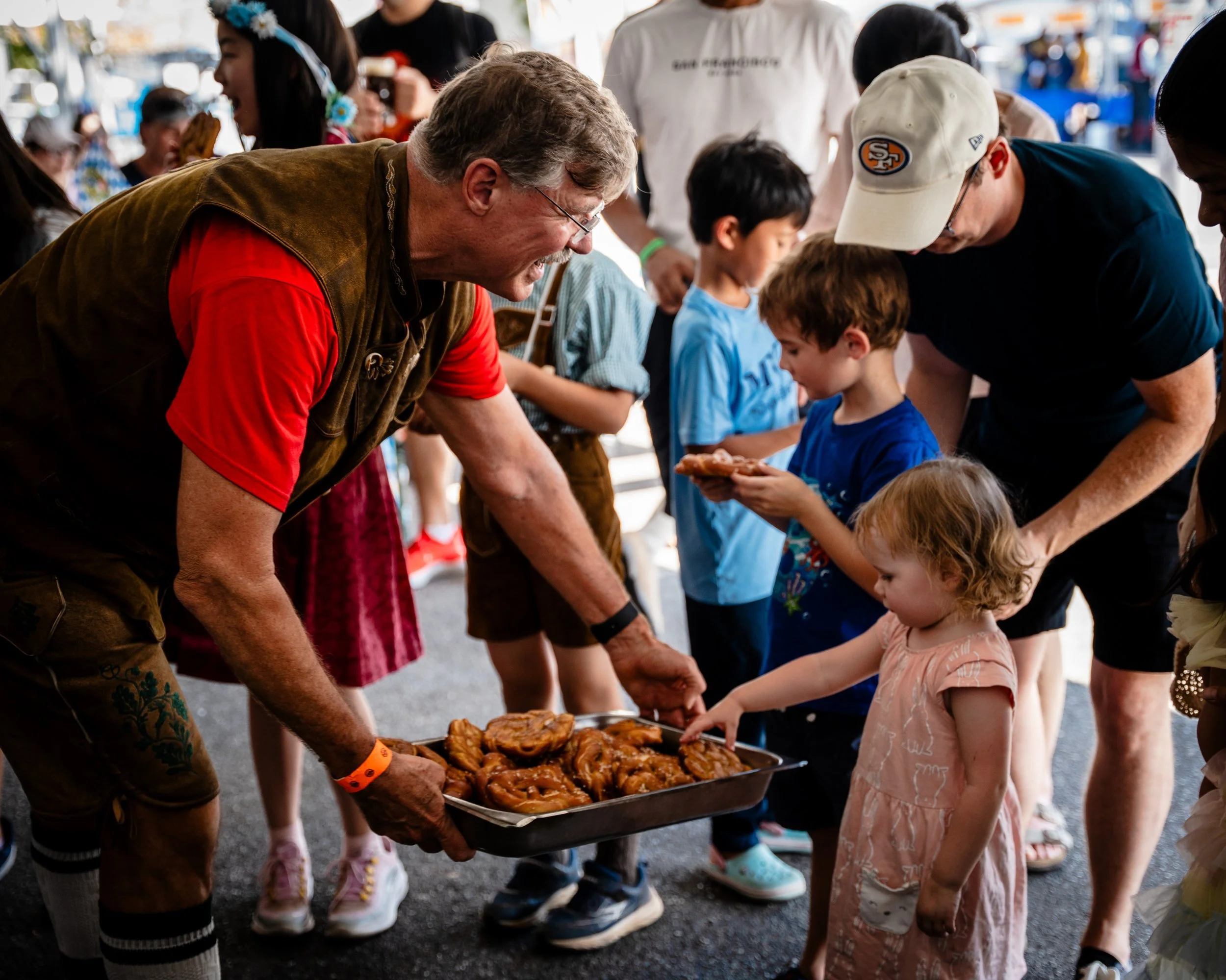 A man dressed in traditional German attire serves pretzels to children at an outdoor event, with other children and adults in the background.