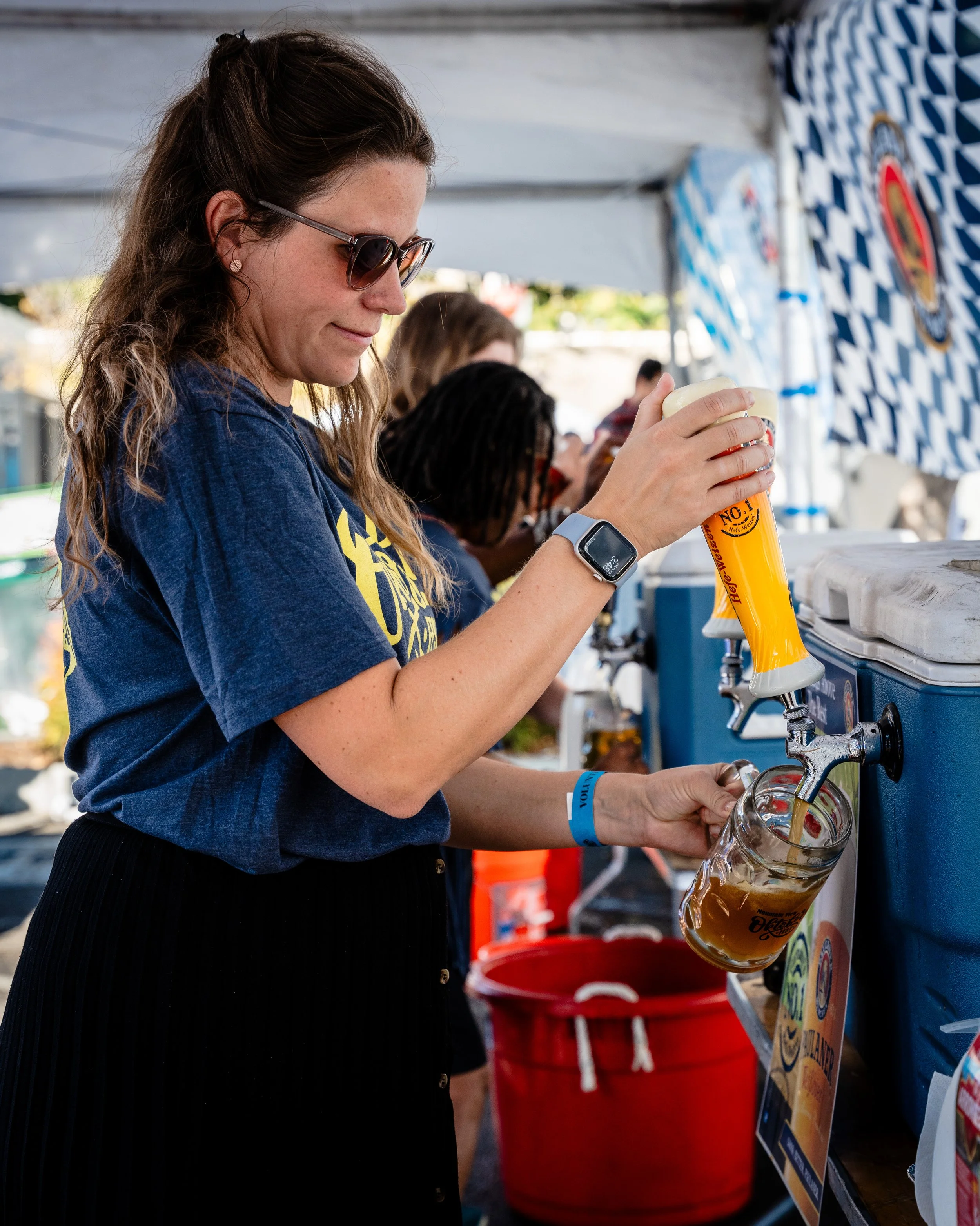 A woman wearing sunglasses and a smartwatch pours beer from a tap into a glass mug at a beer festival or outdoor event.
