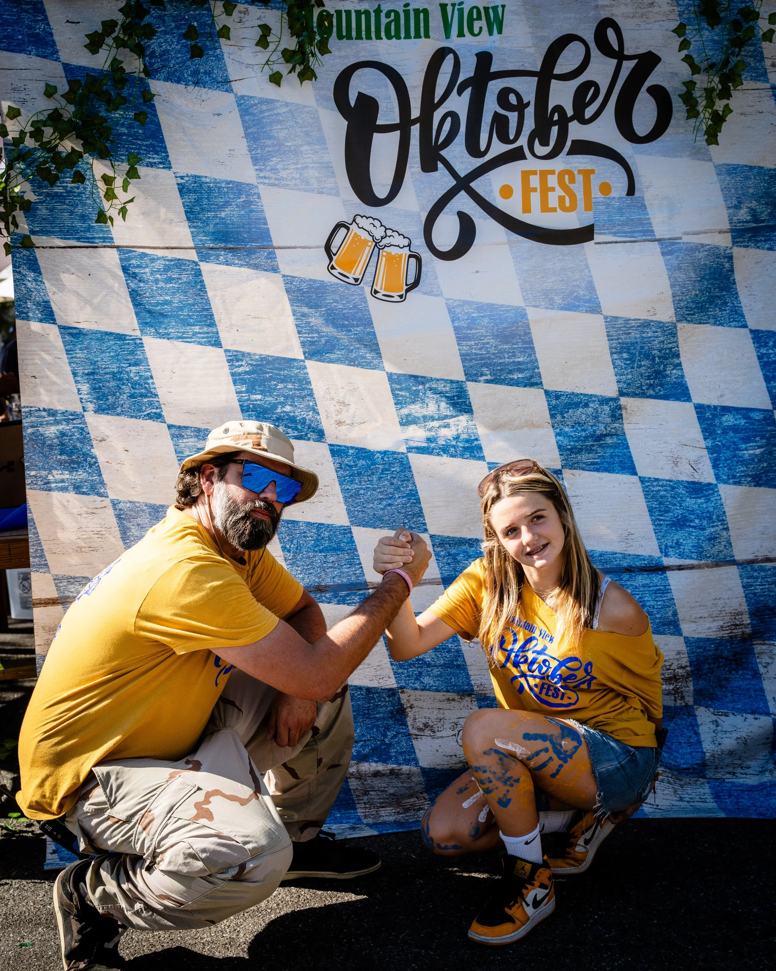 A man and a girl are posing at the Oktoberfest event, shown crouching down and flexing their arm muscles while doing a strength contest. The backdrop features blue and white checkered pattern with the text 'Mountain View Oktoberfest' and beer mug ill