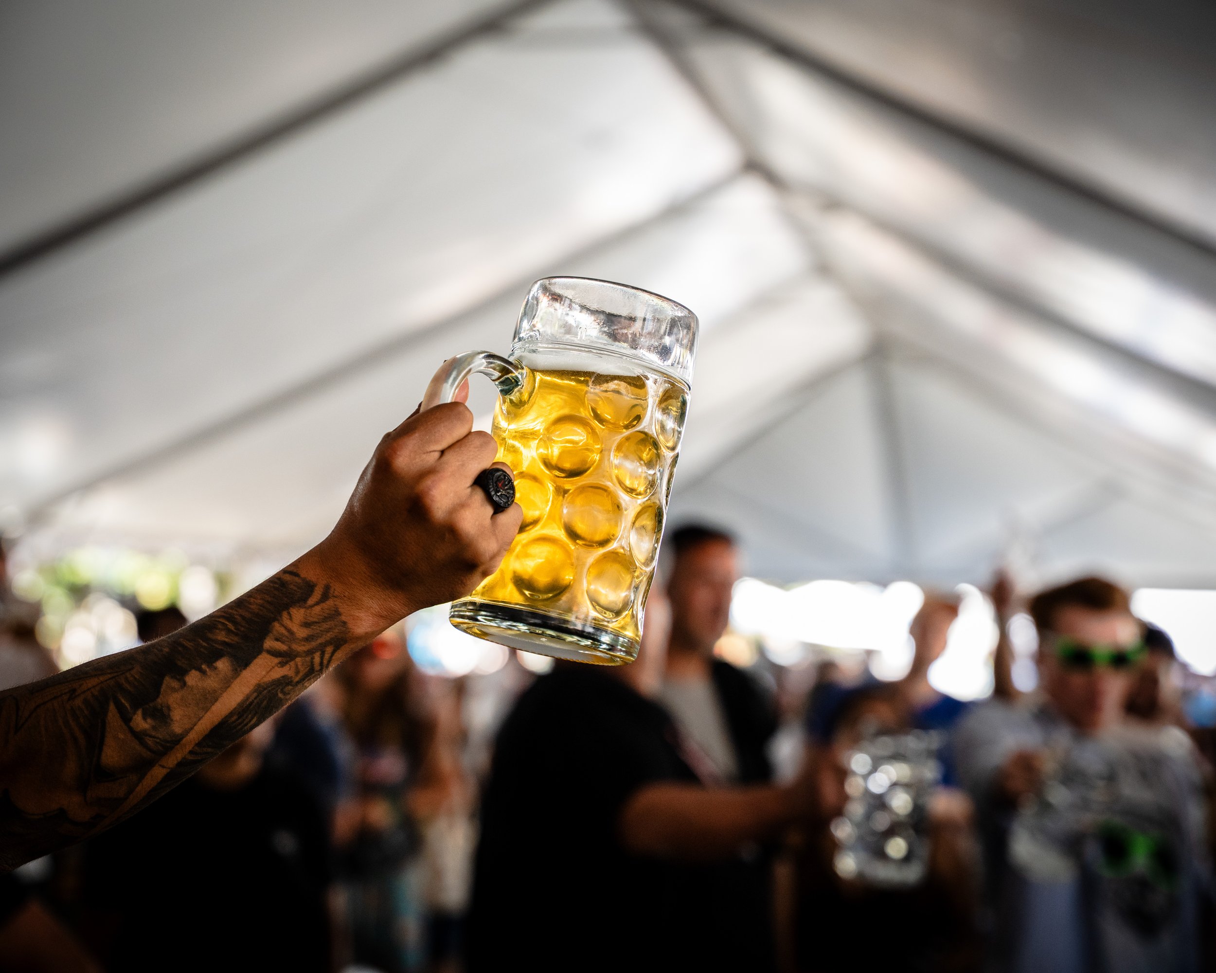 Close-up of a tattooed hand holding a large mug of beer at a lively gathering under a white tent.