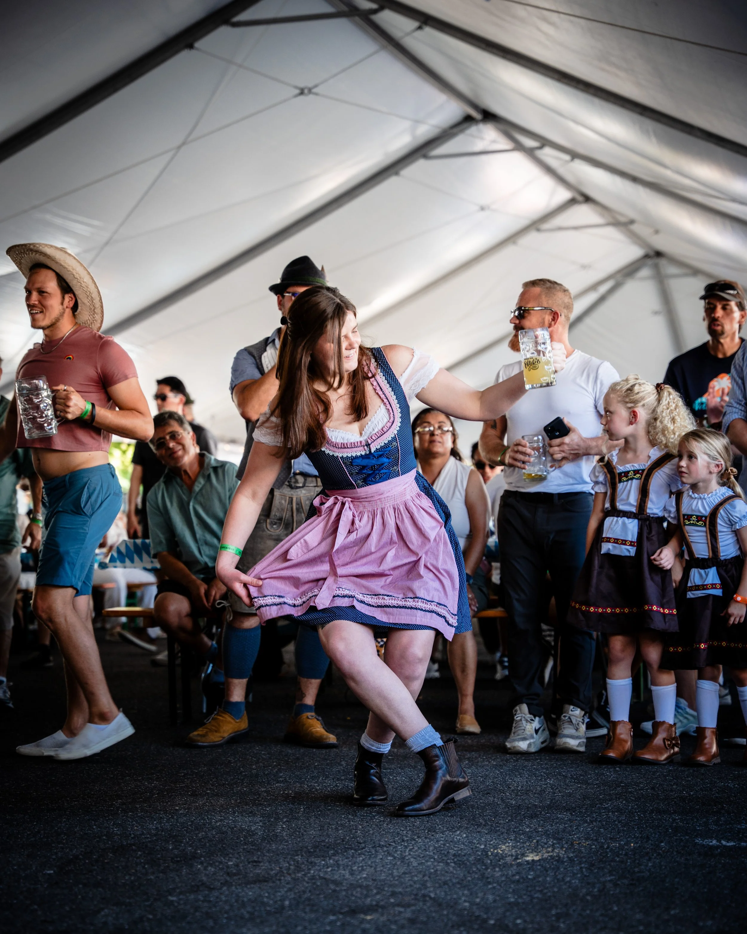 Group of people enjoying a traditional beer festival inside a large tent, with some dressed in traditional German attire, dancing and holding beer steins.