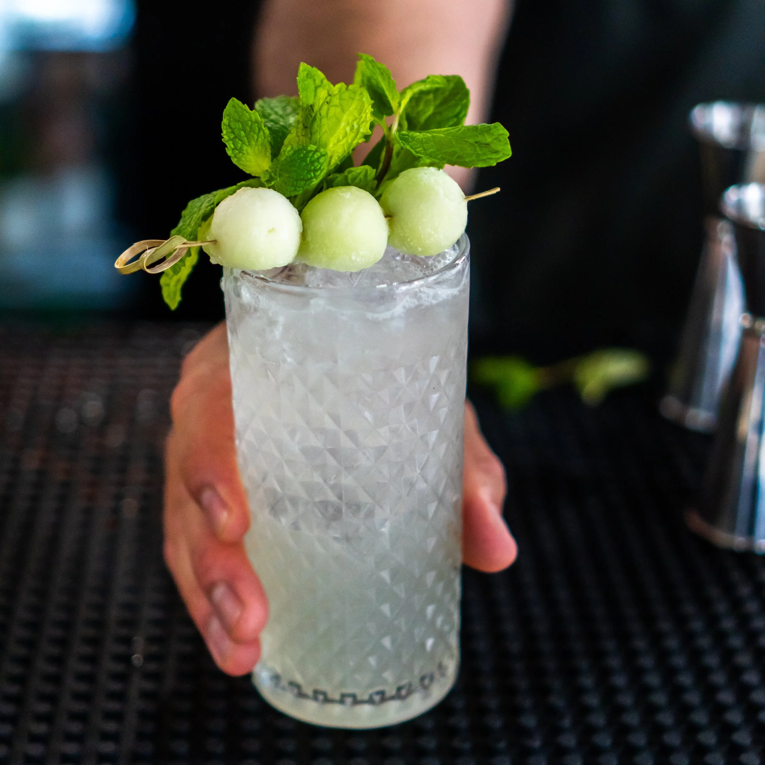 A cocktail garnished with mint leaves and melon balls on a skewer, served over ice in a tall glass at bar in Maryland.