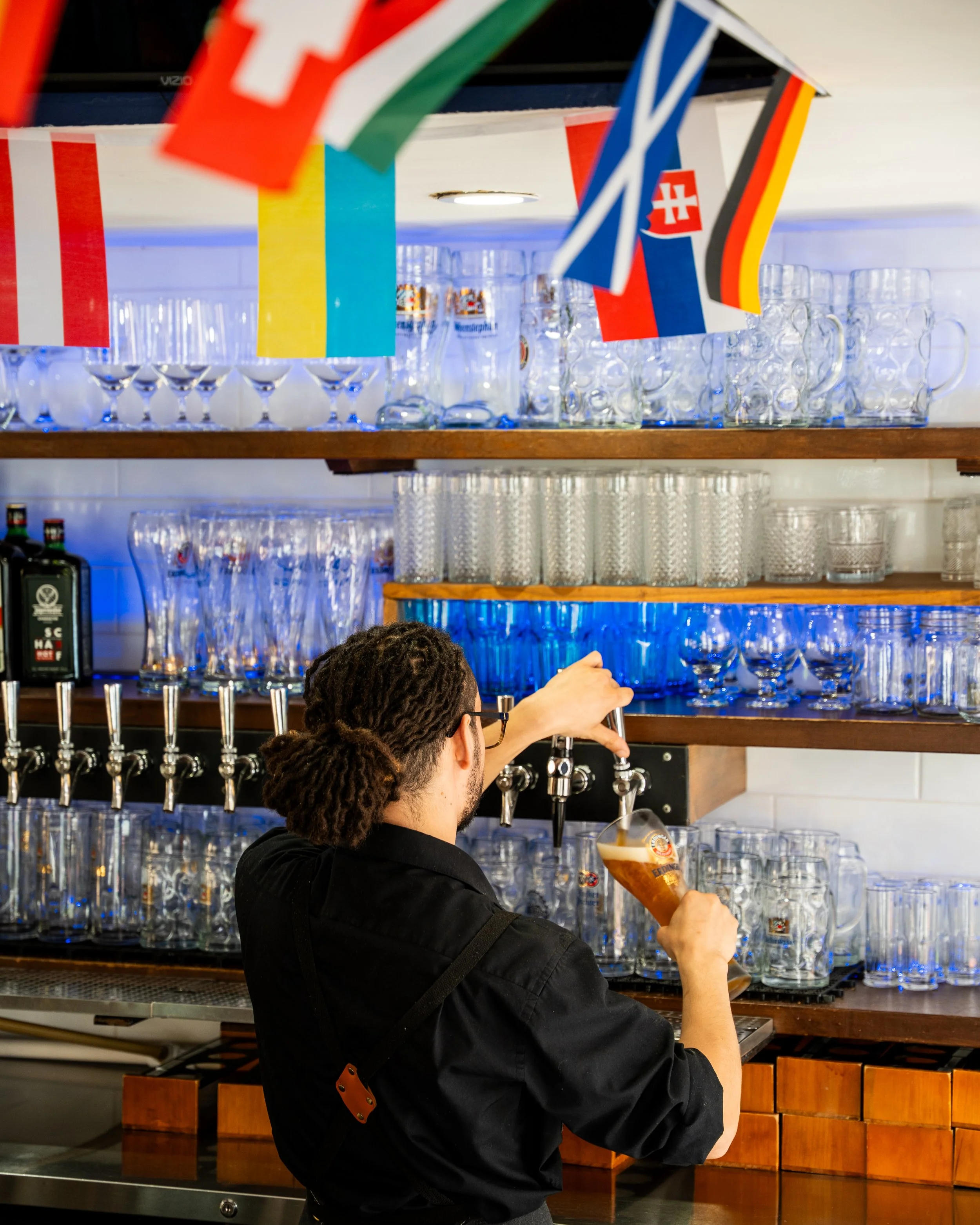 Bartender pouring a beer from a tap behind a bar with shelves of glassware and international flags hanging above.