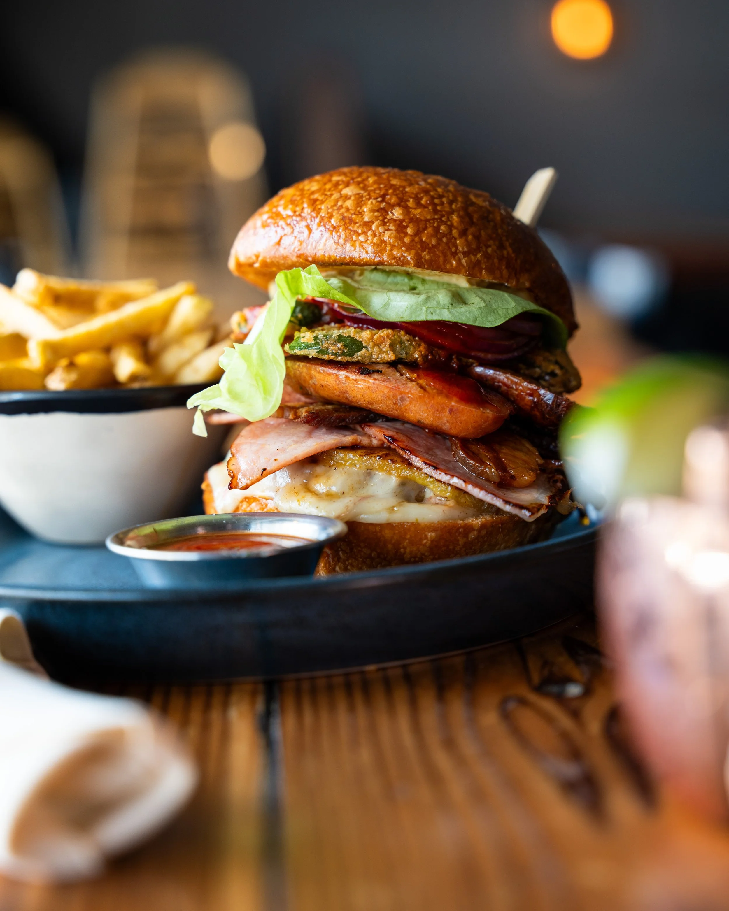 A tall burger with lettuce, tomato, bacon, cheese, and multiple patties, served with French fries and a small side of ketchup.