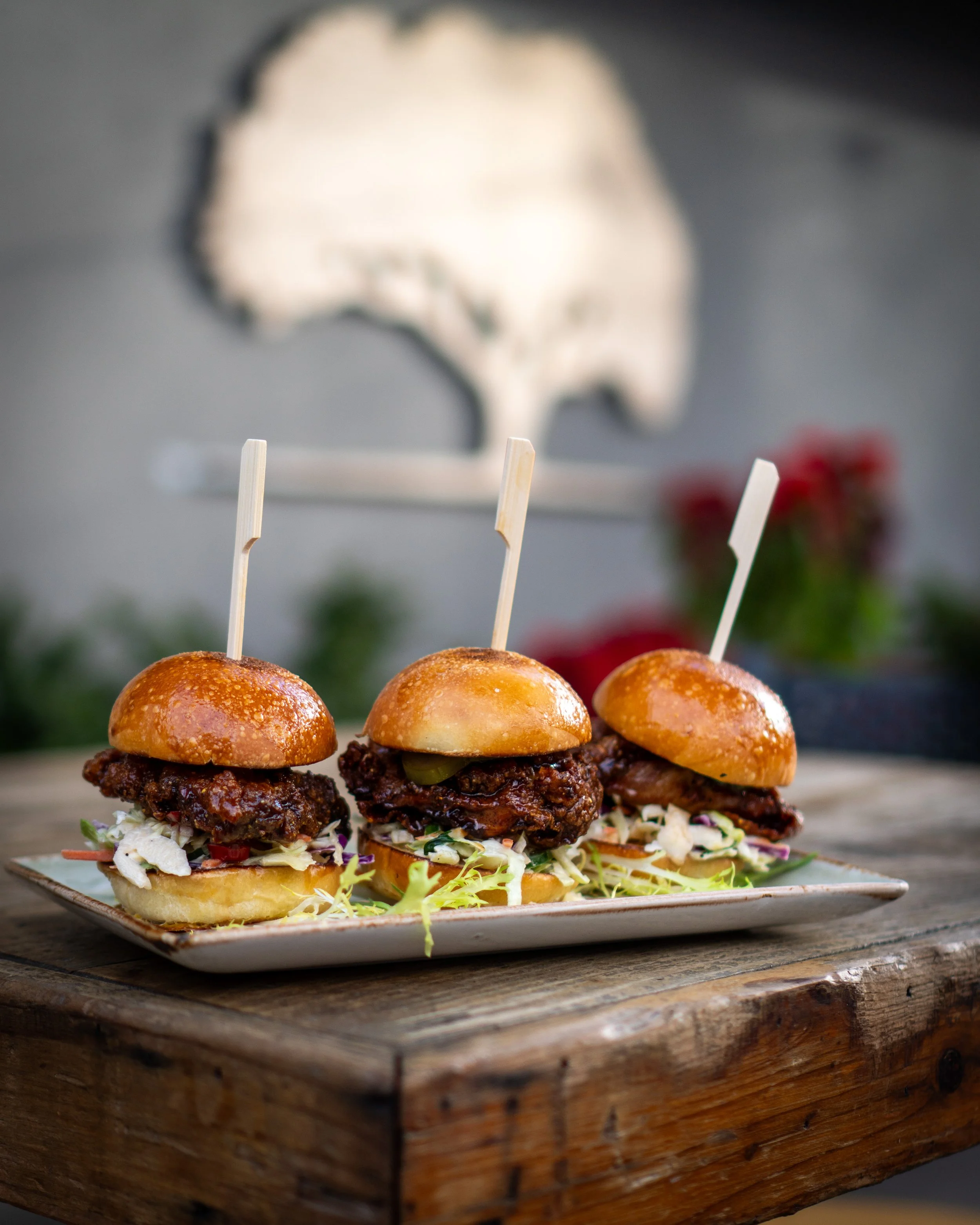 Three small barbecue sliders with coleslaw on white plates, on a rustic wooden table, with a blurred background that has an outline of Australia.