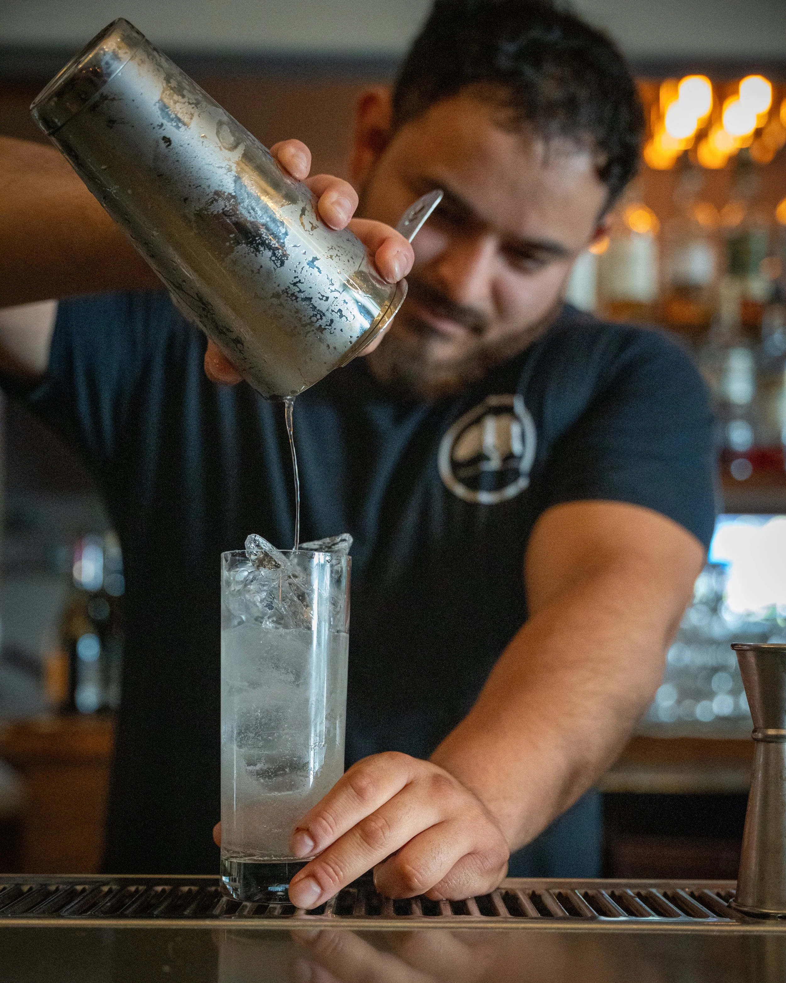 A bartender wearing a black shirt with a circular logo is pouring a liquid from a metal cocktail shaker into a tall glass filled with ice.