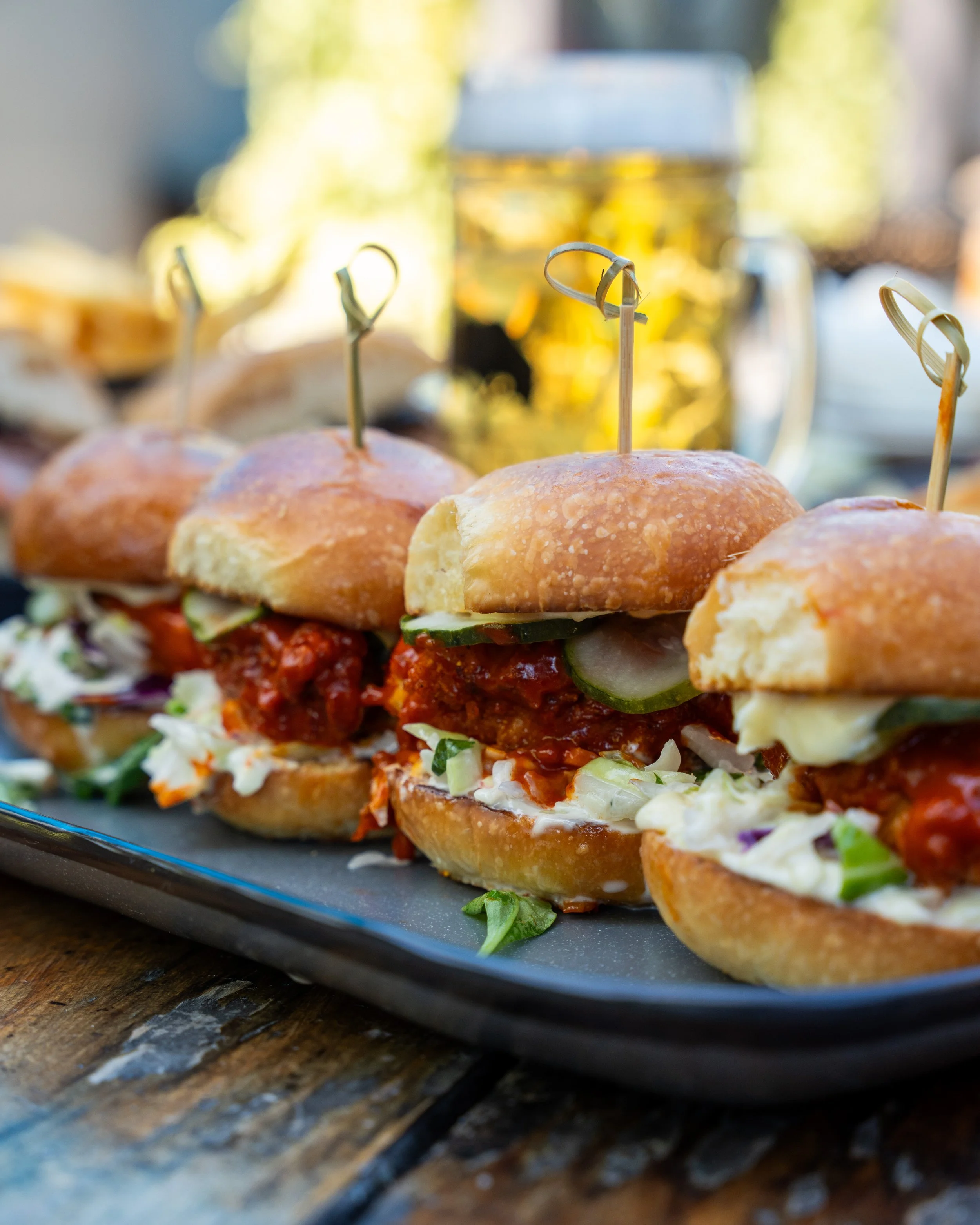 Three sliders with pickles, coleslaw, and sauce on a black tray, with a mug of beer in the background.
