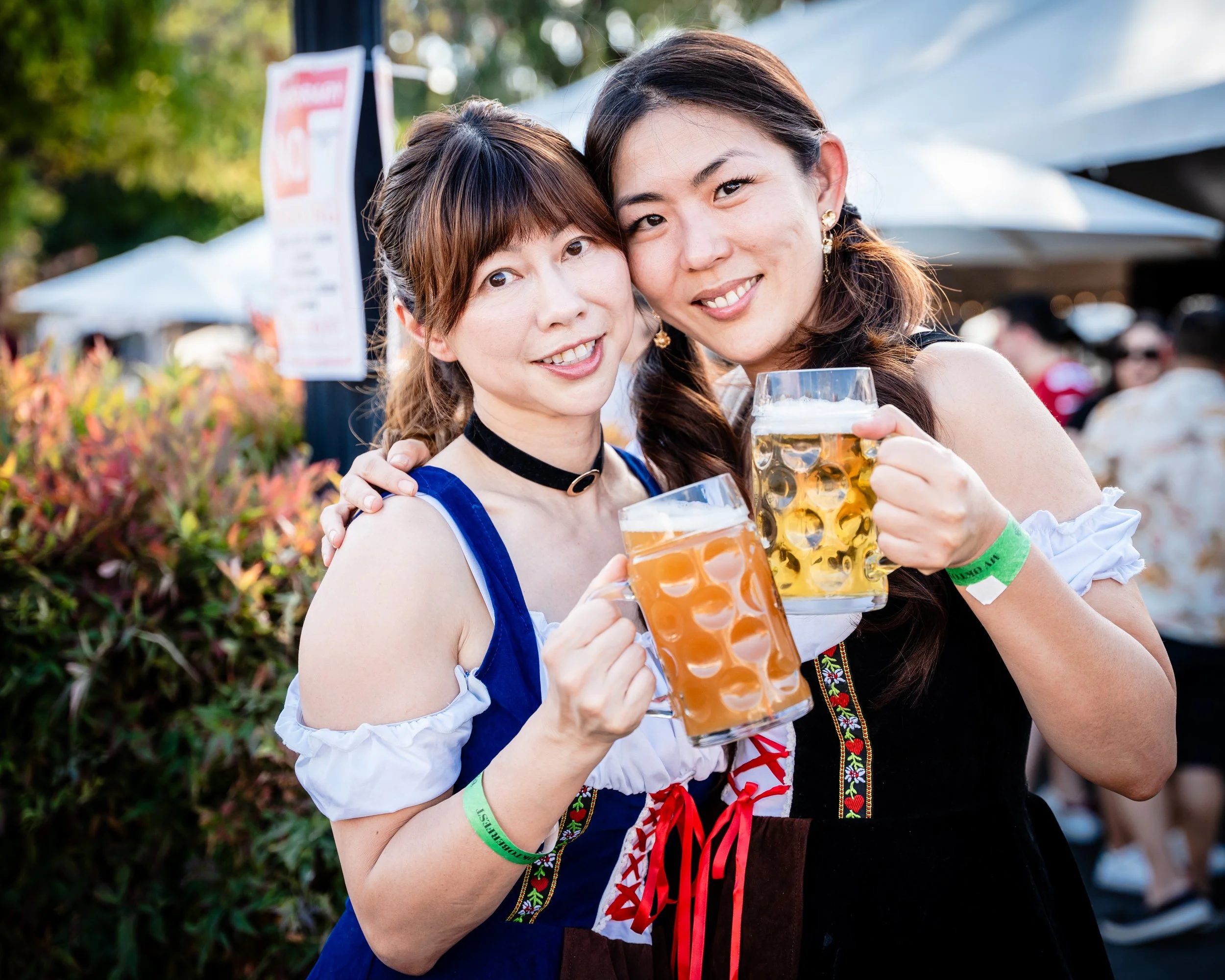 Two women wearing traditional European dirndl dresses celebrating outdoors, holding large glasses of beer, smiling, with festival tents and a crowd in the background.