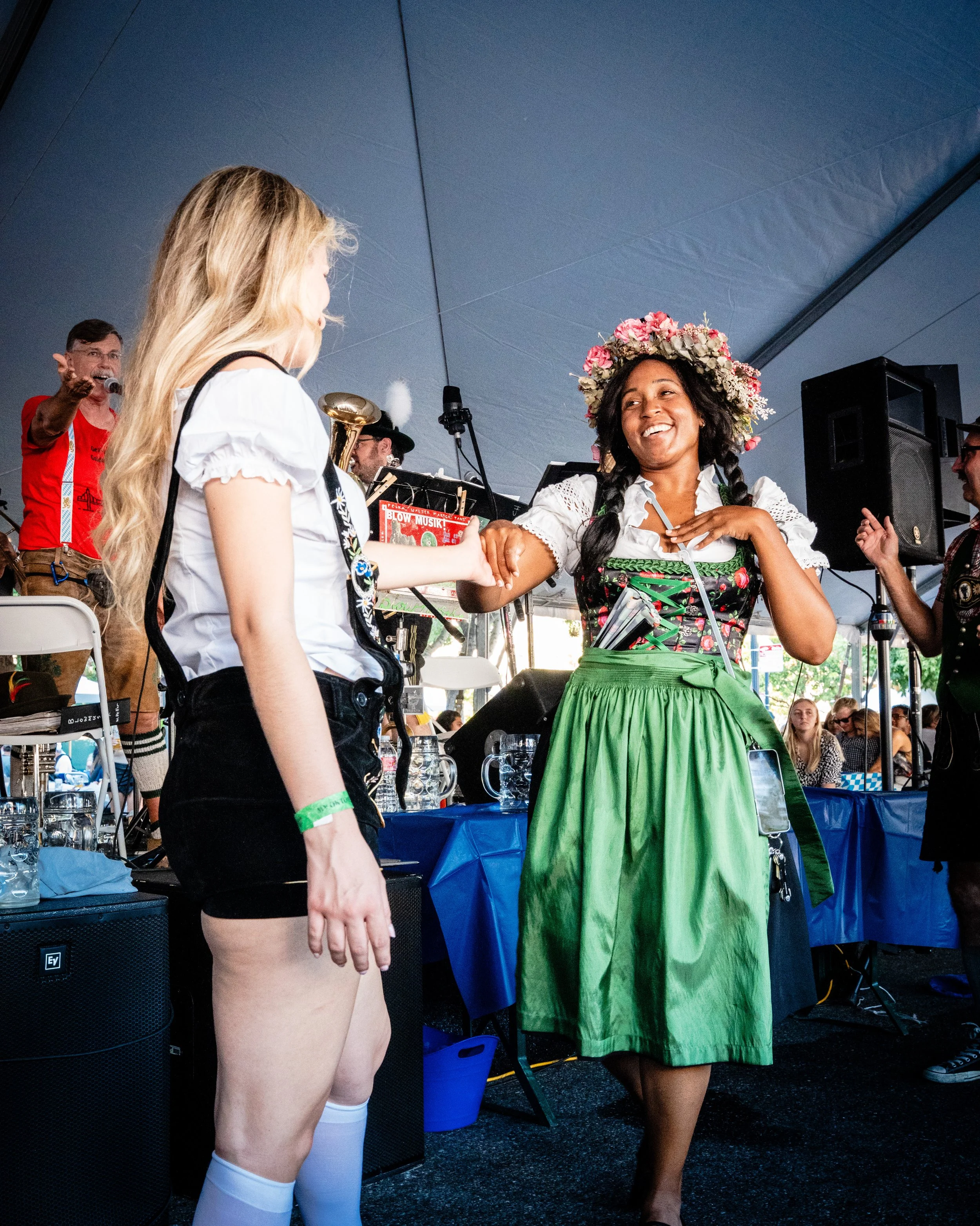Two women shaking hands on stage at a festival. The woman on the right wears a flower crown and traditional dress, while the woman on the left wears a white shirt and black shorts. There is a band playing in the background.