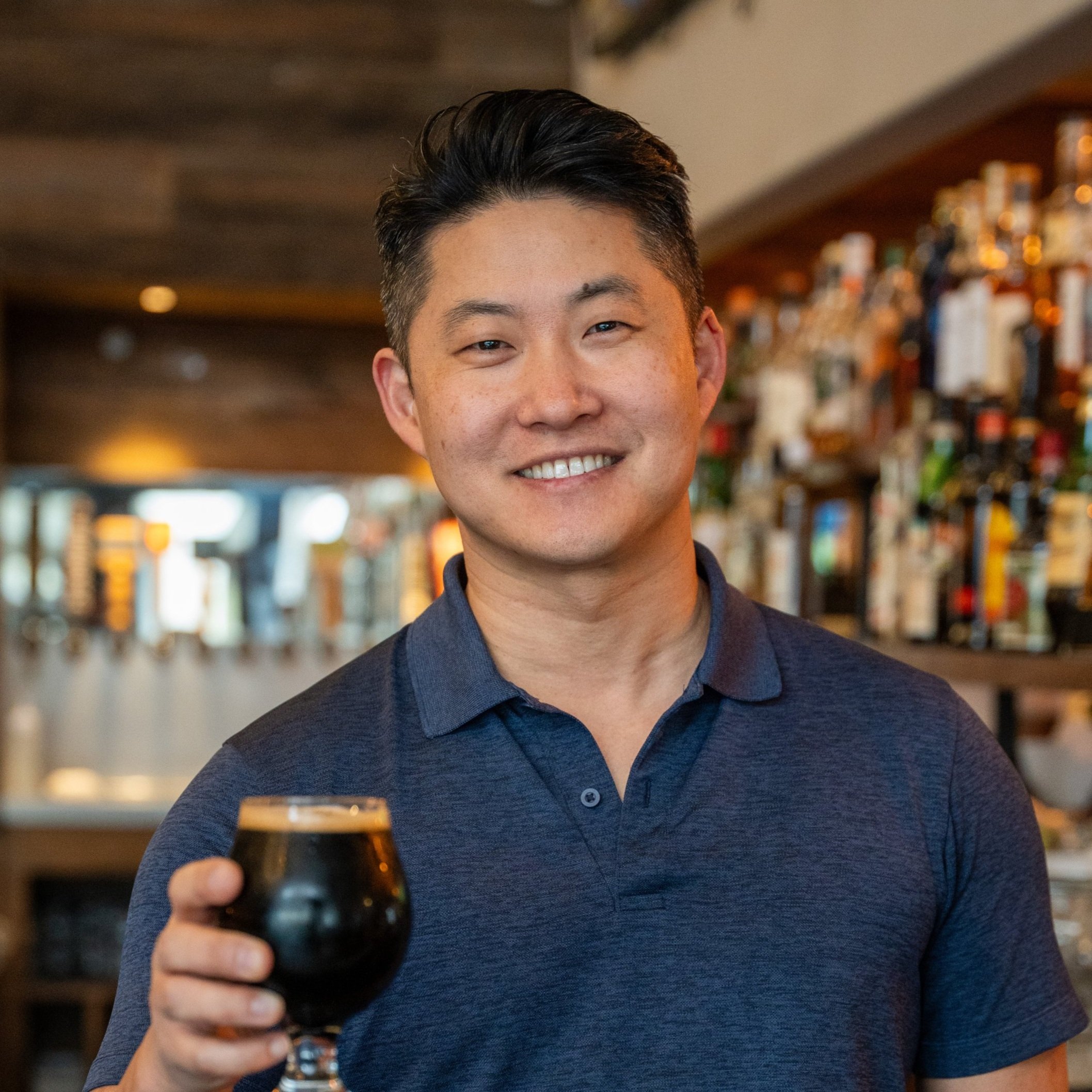 A smiling man holding a glass of dark beer in a bar with a blurred background of bottles on shelves.