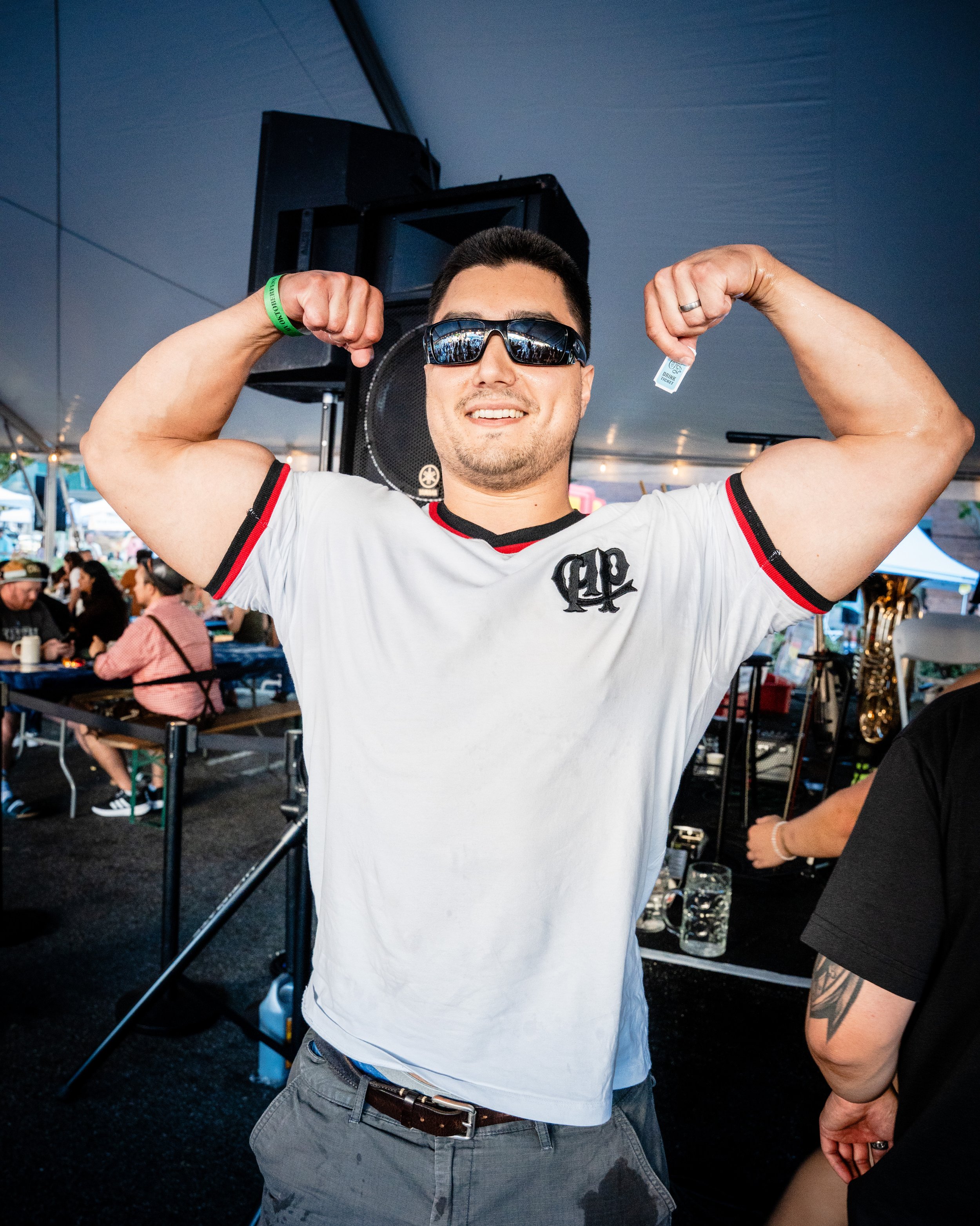 A man smiling and flexing muscles, wearing sunglasses and a white t-shirt, at a gathering under a large tent.
